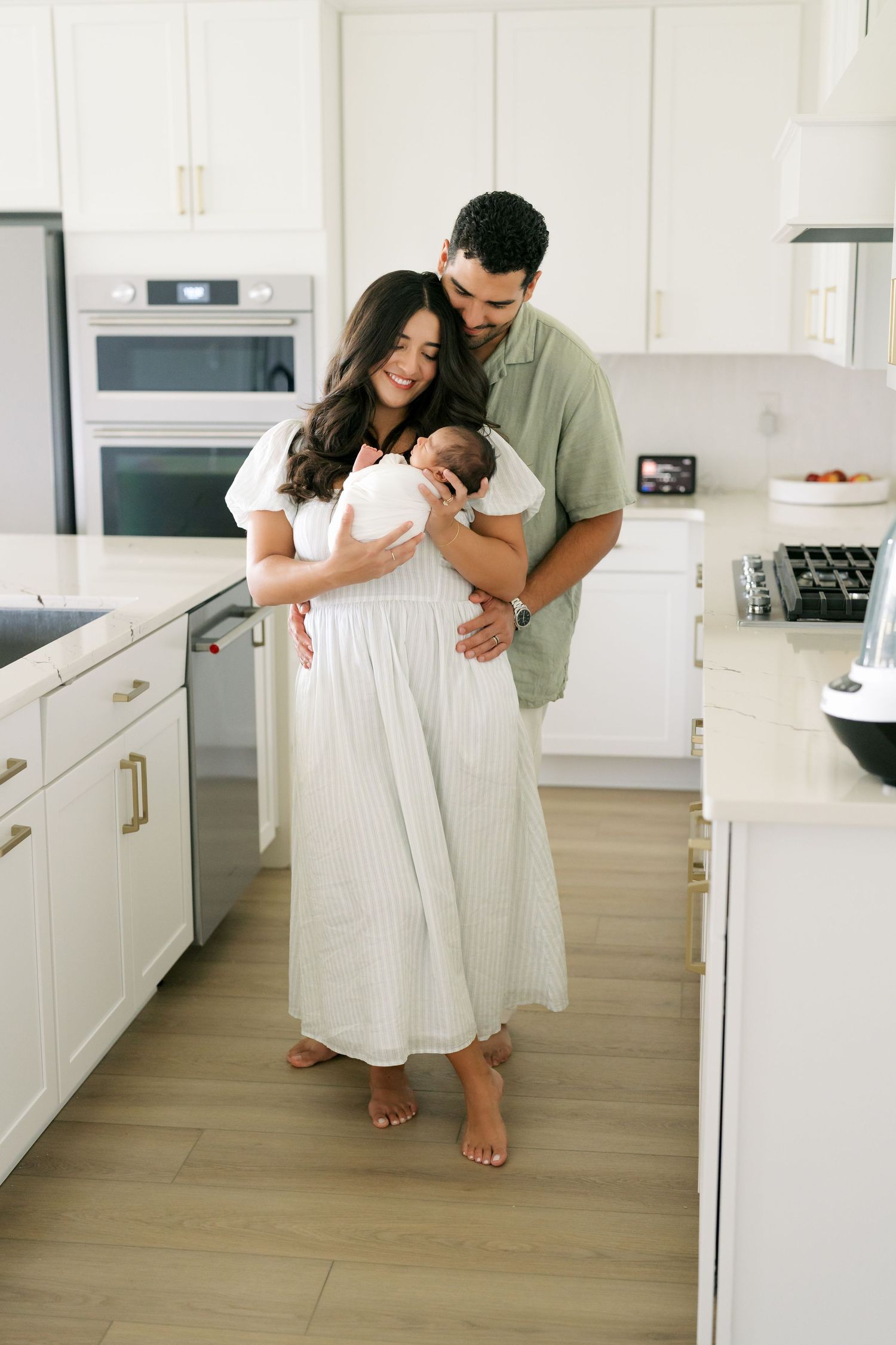 A couple in a bright white kitchen lovingly gazes at their newborn baby, the mother in a white dress.