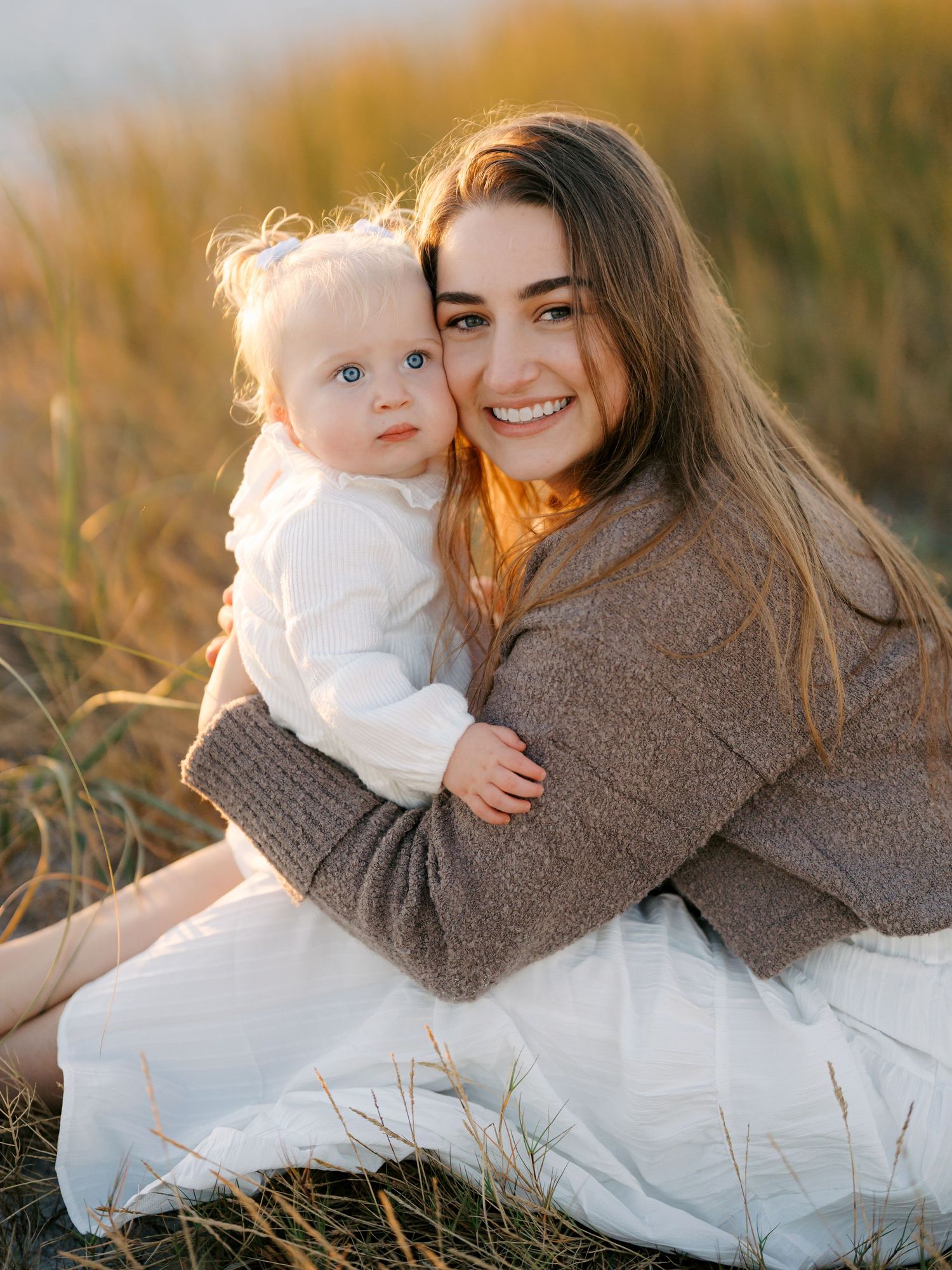 Mother and baby share tender moments in warm sunset light while wearing cozy sweaters on grassy field.