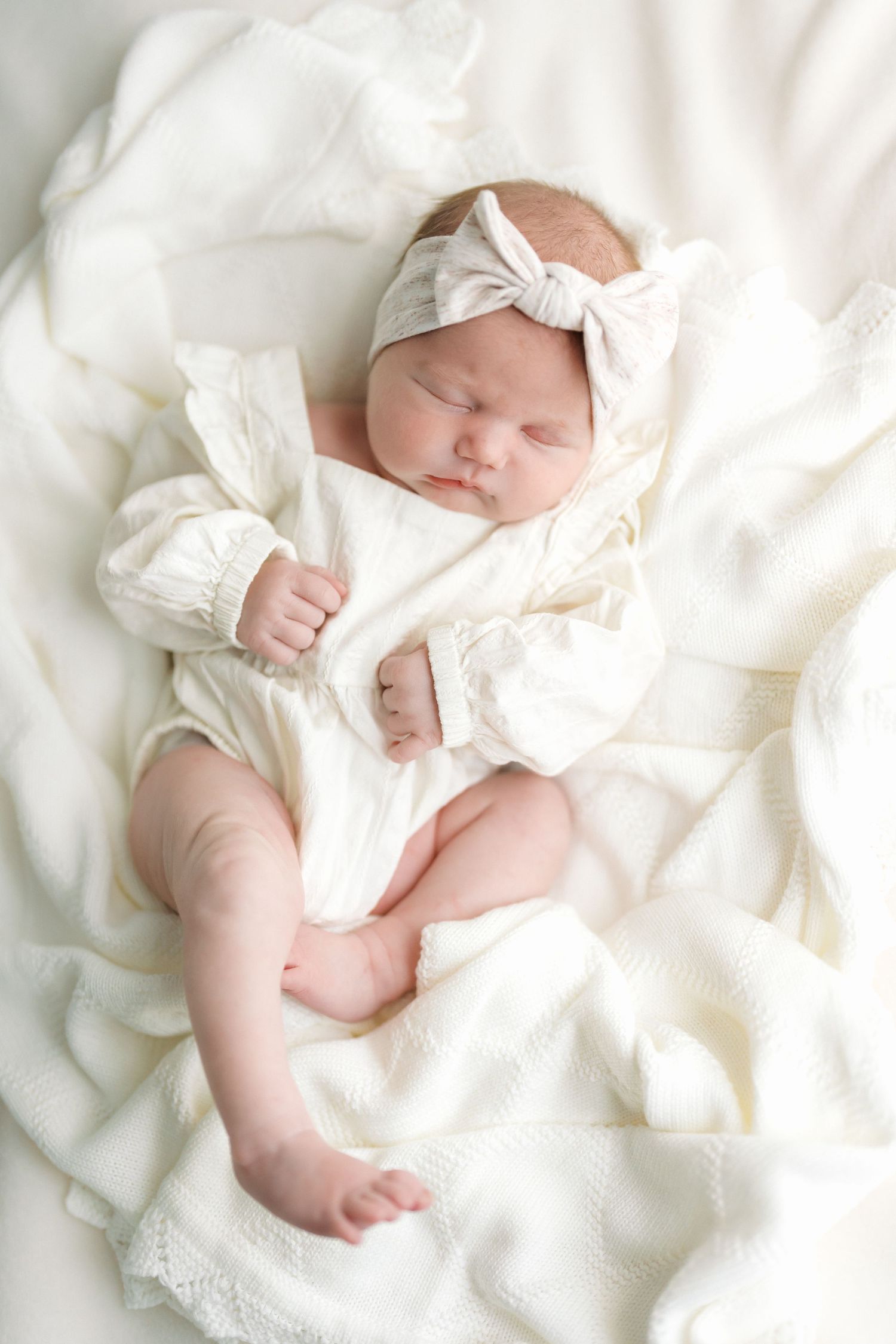 Peaceful newborn baby girl sleeping in white outfit and bow headband on soft white blanket.