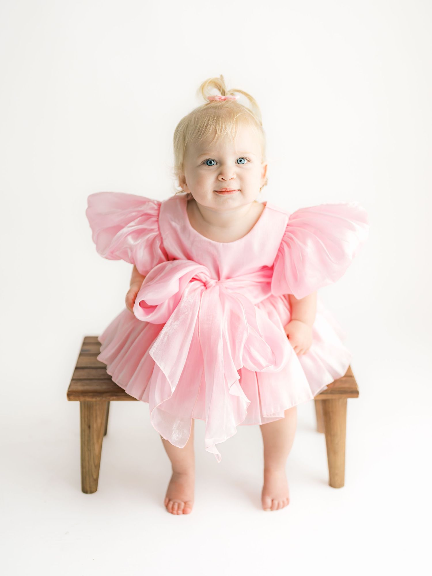 A toddler in a pink tulle dress sits and plays on a wooden bench against a white background.