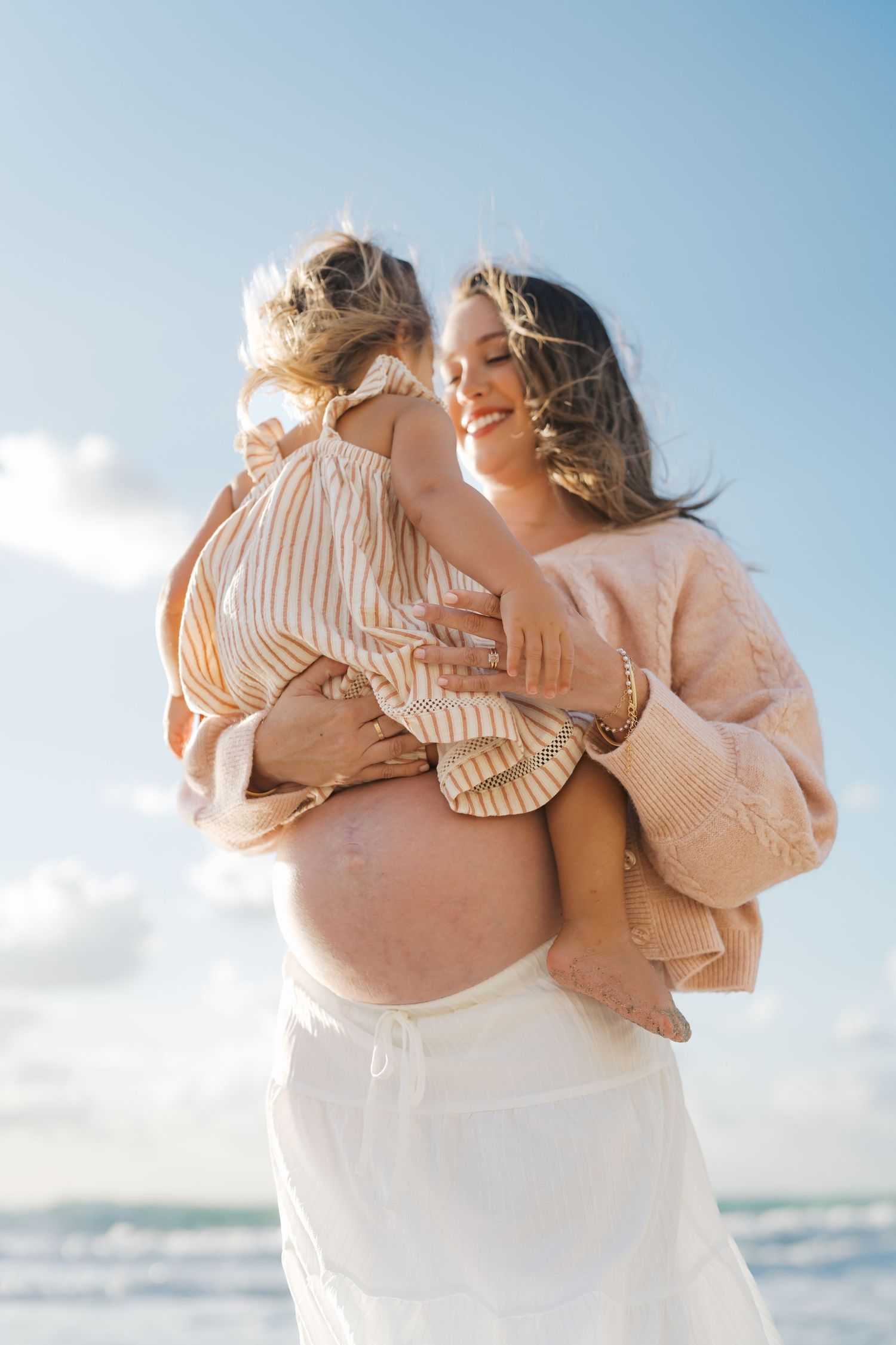 Pregnant woman in white skirt holds young child at beach, smiling under blue sky.