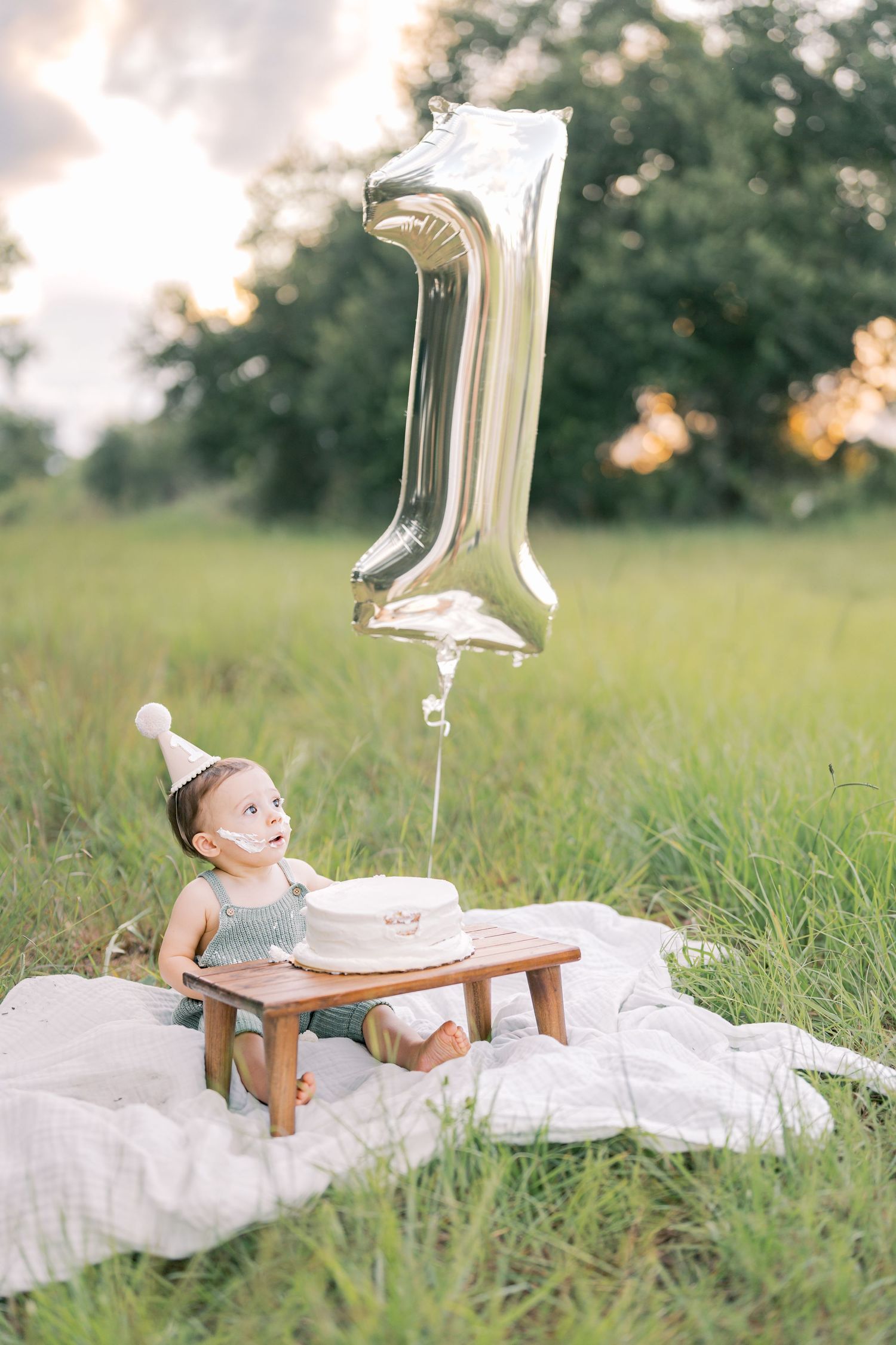 A baby in white outfit sits on a wooden bench with a silver number 1 balloon in an outdoor grassy setting.