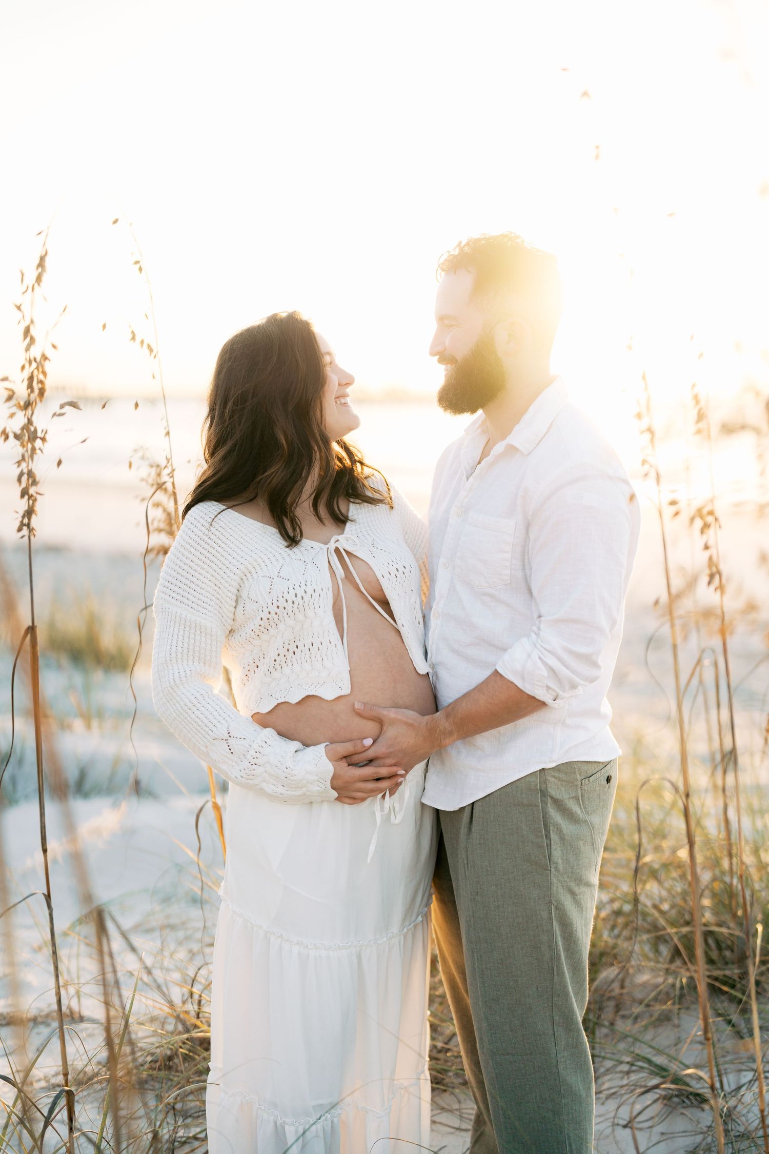Pregnant couple embracing on a sunlit beach, woman in white dress, man in white shirt and green pants.