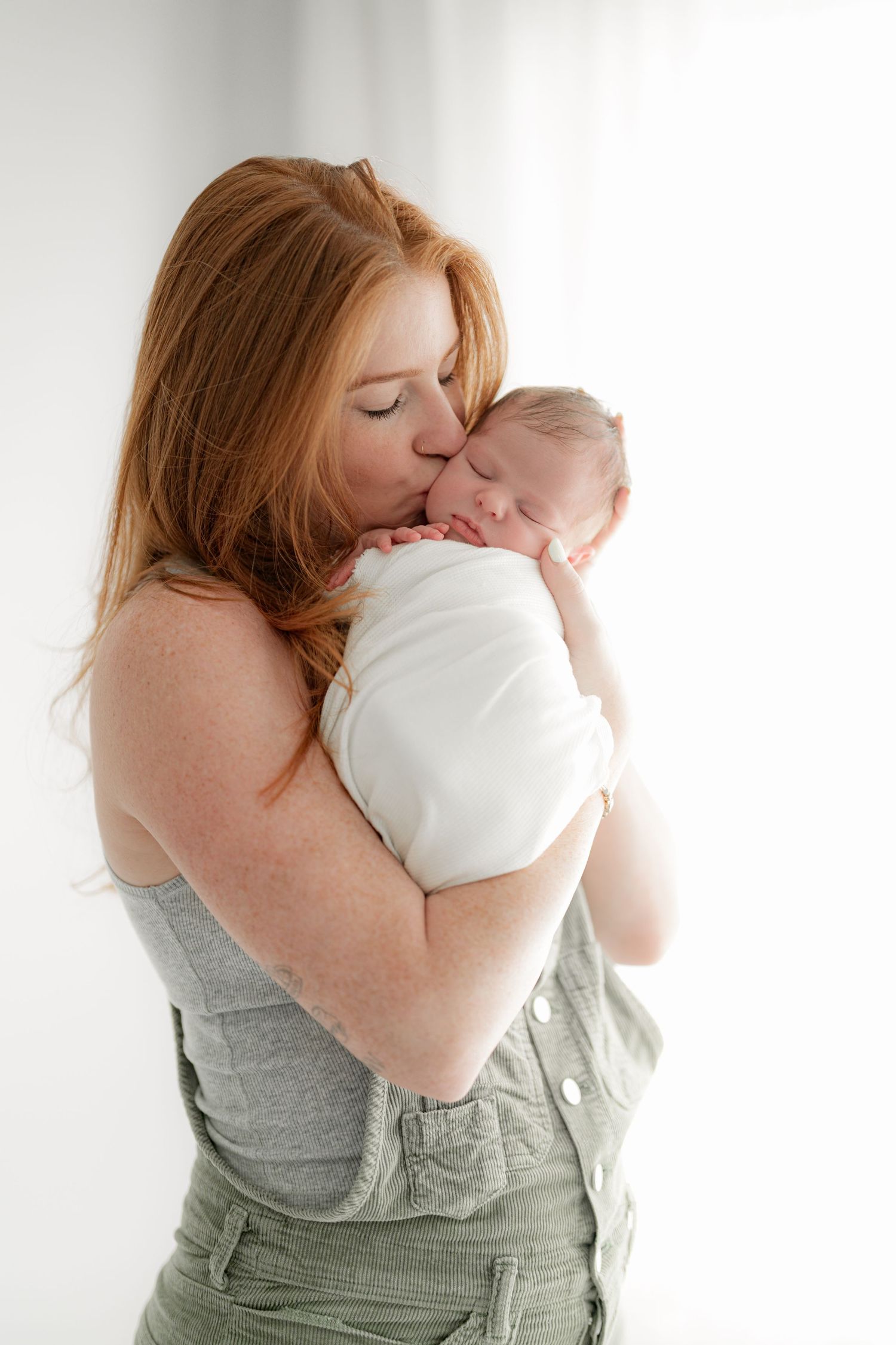 A woman in grey attire tenderly kisses a swaddled sleeping newborn.