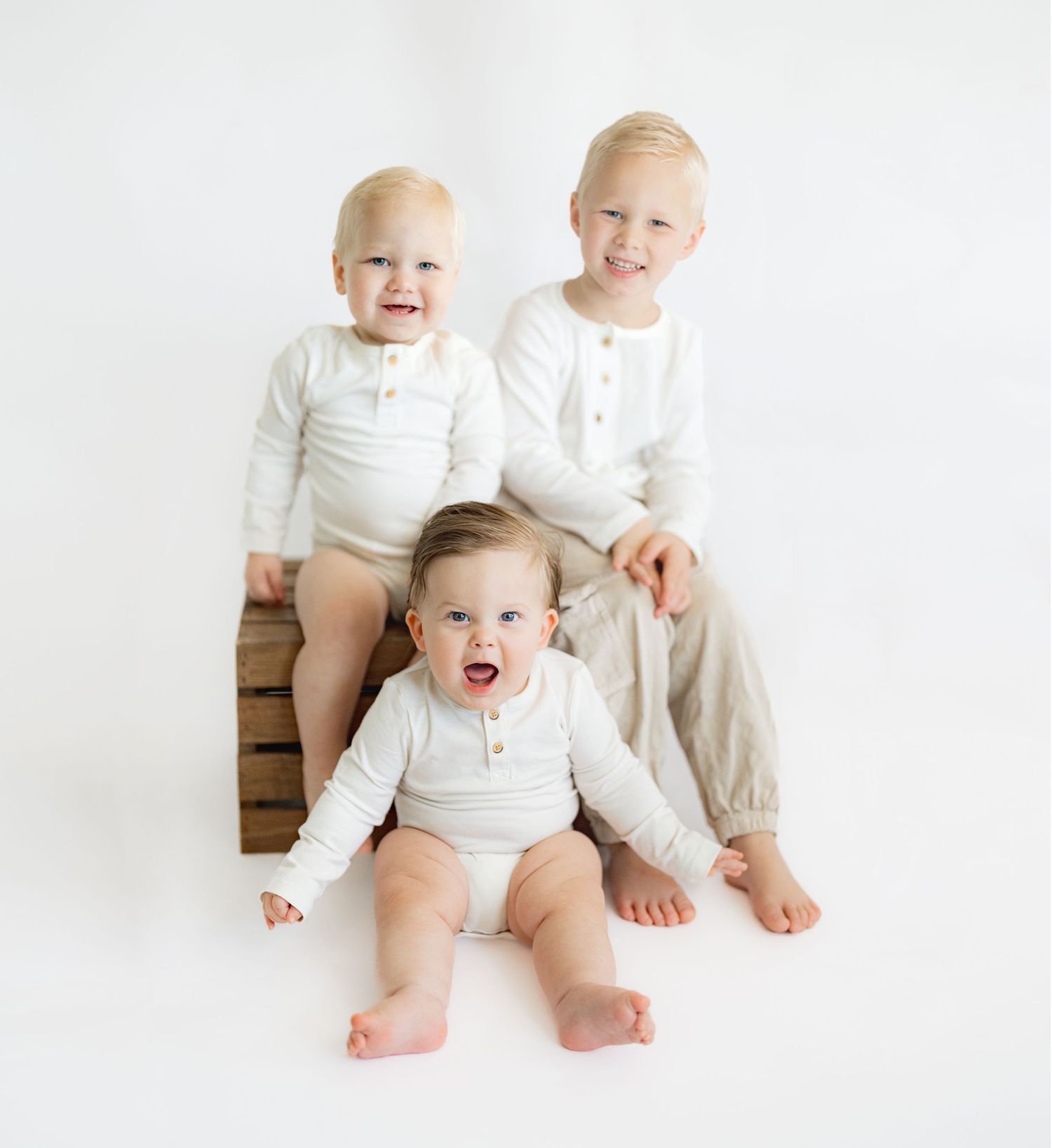 Three babies wearing white outfits sit together on a wooden bench against a white backdrop.