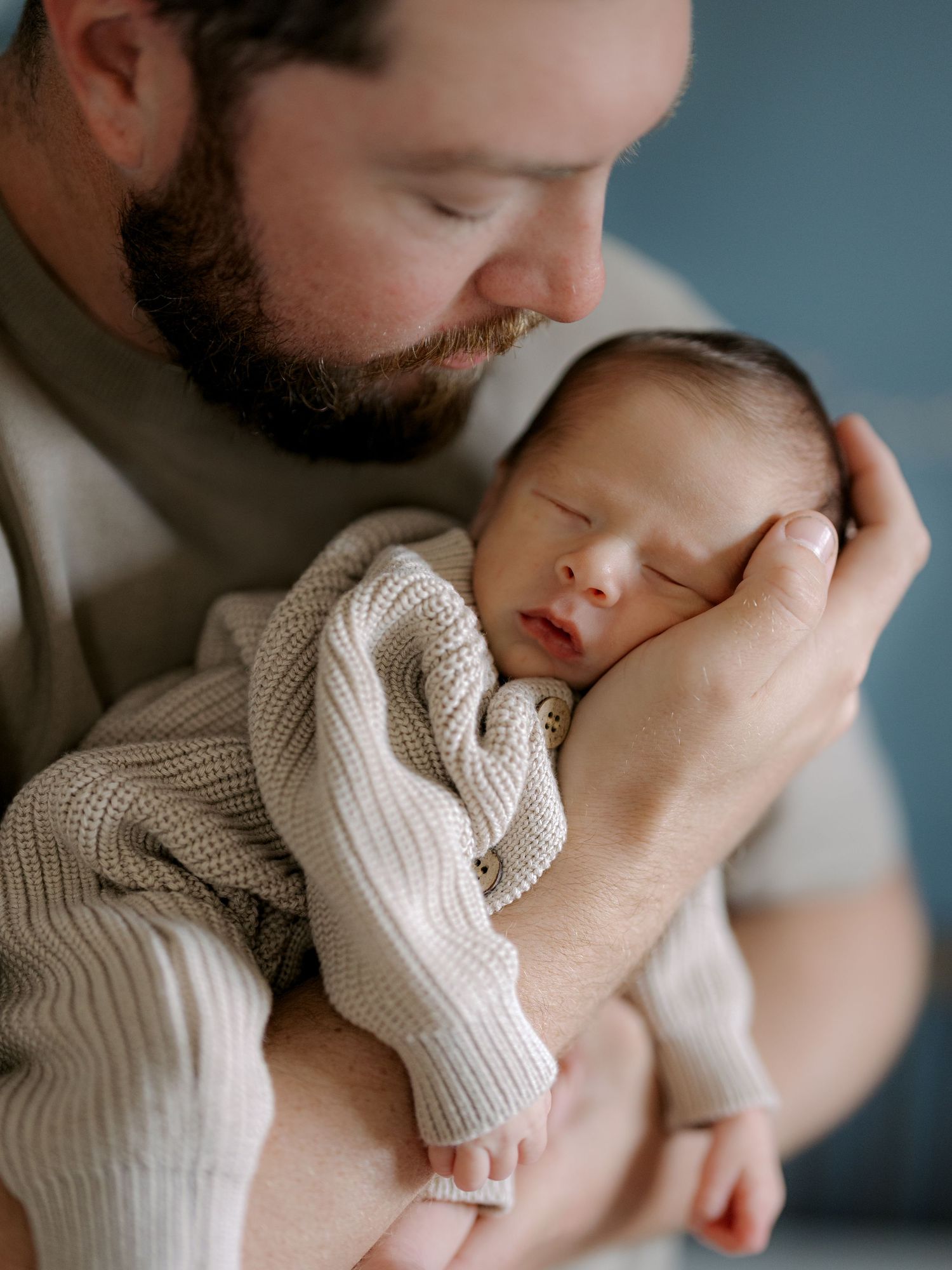 A bearded father tenderly holds his sleeping newborn baby dressed in a cream knit outfit.