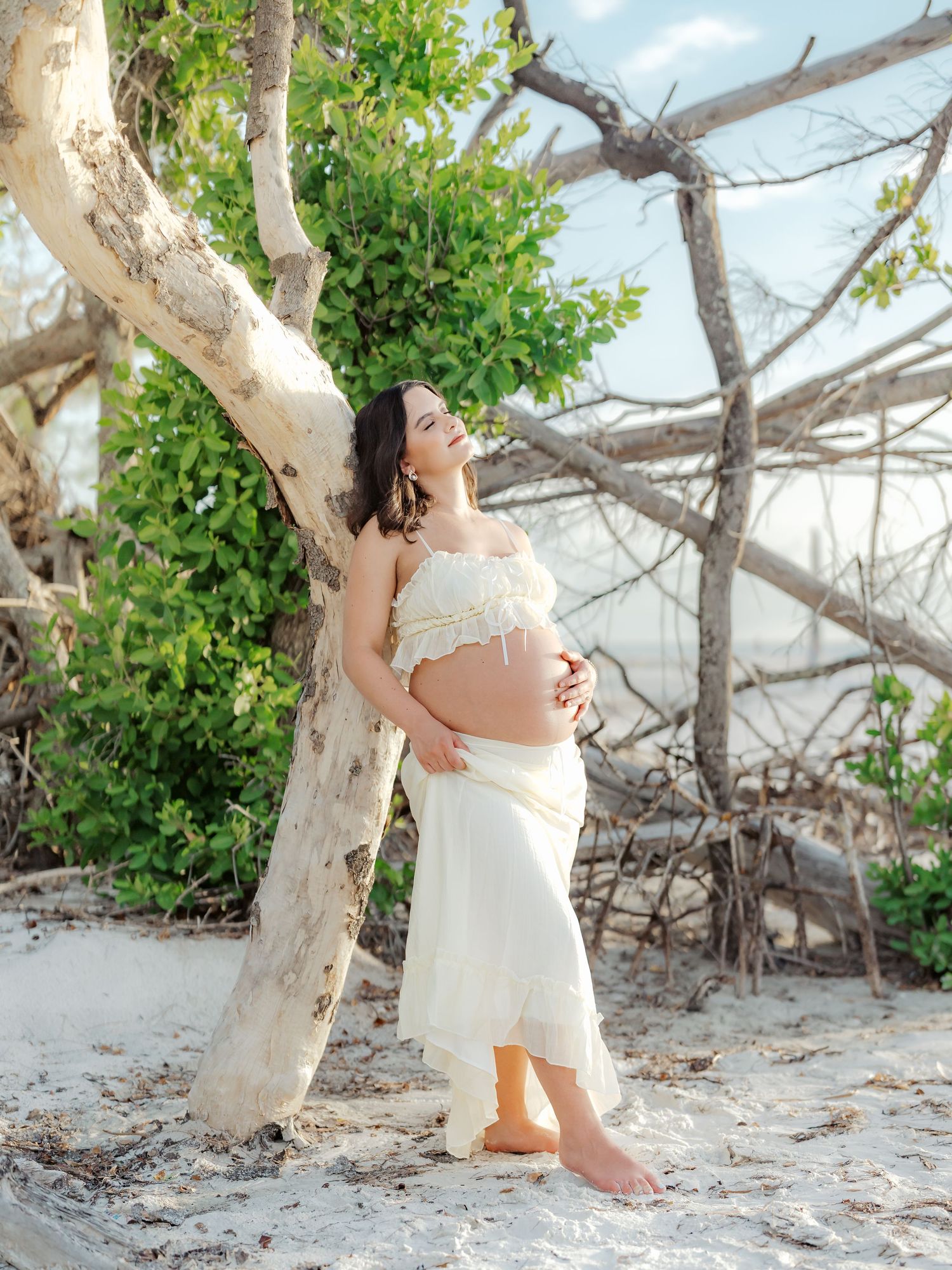 A romantic beach scene with a couple posing near white tree trunks and sand during sunset.