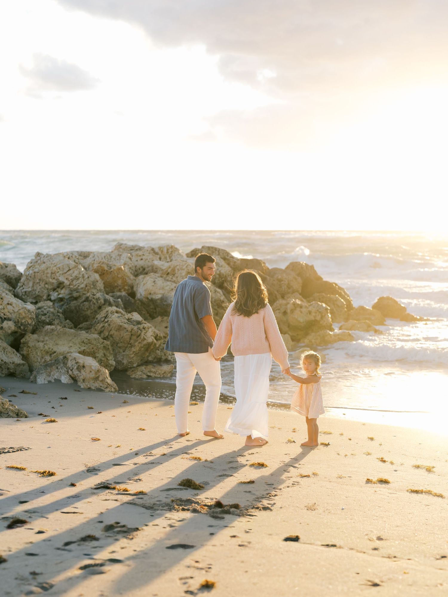 Family of three holding hands on a sandy beach at golden sunset with rocky shoreline in the background.