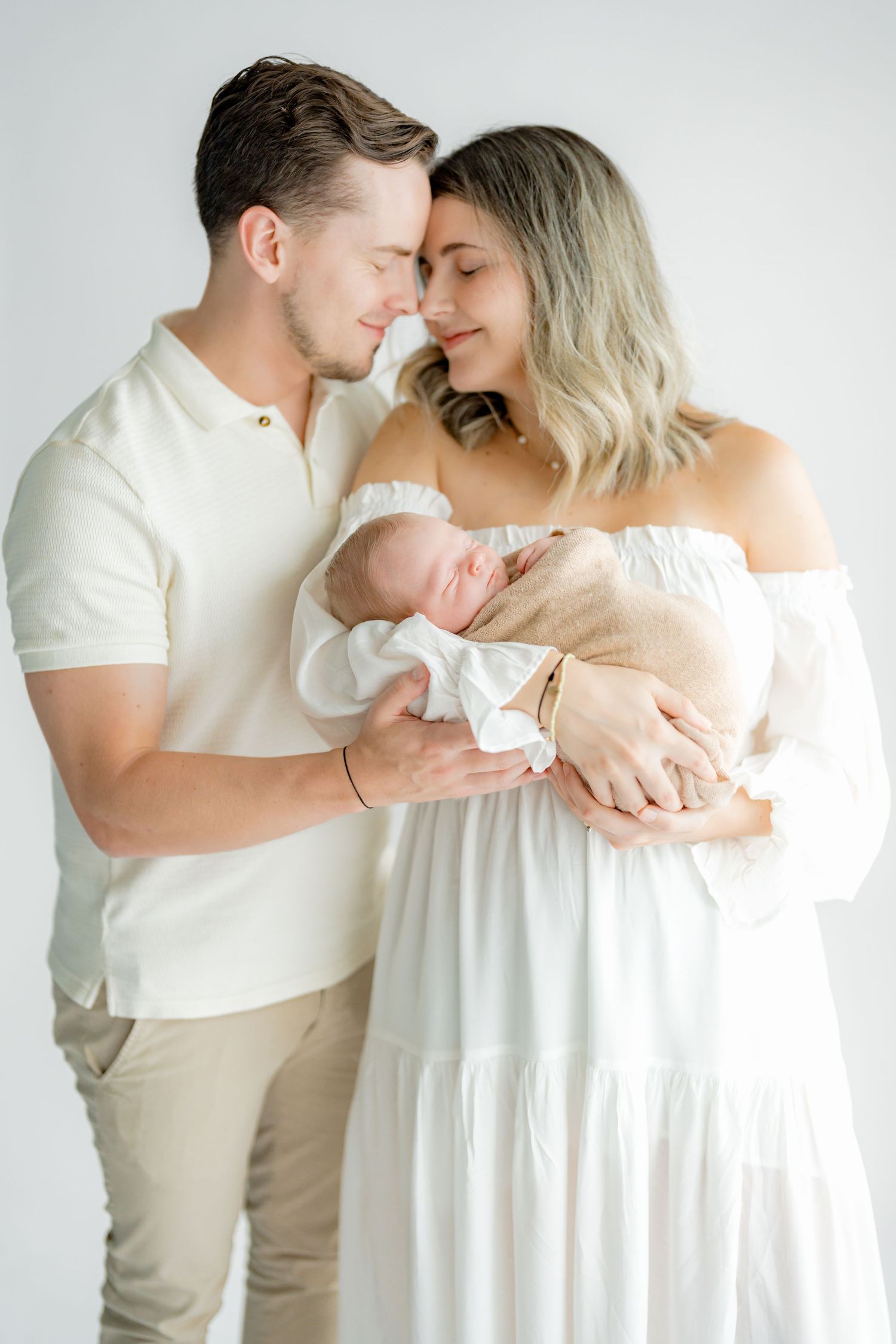A couple in neutral tones tenderly holds their sleeping newborn baby in a bright white studio setting.