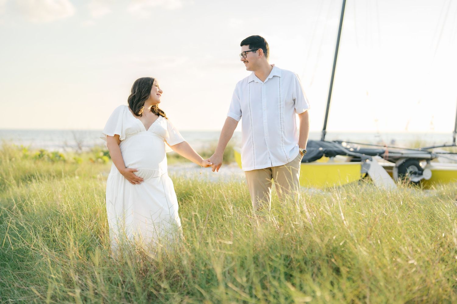 Couple walking hand in hand through a sunlit meadow wearing white outfits during golden hour.