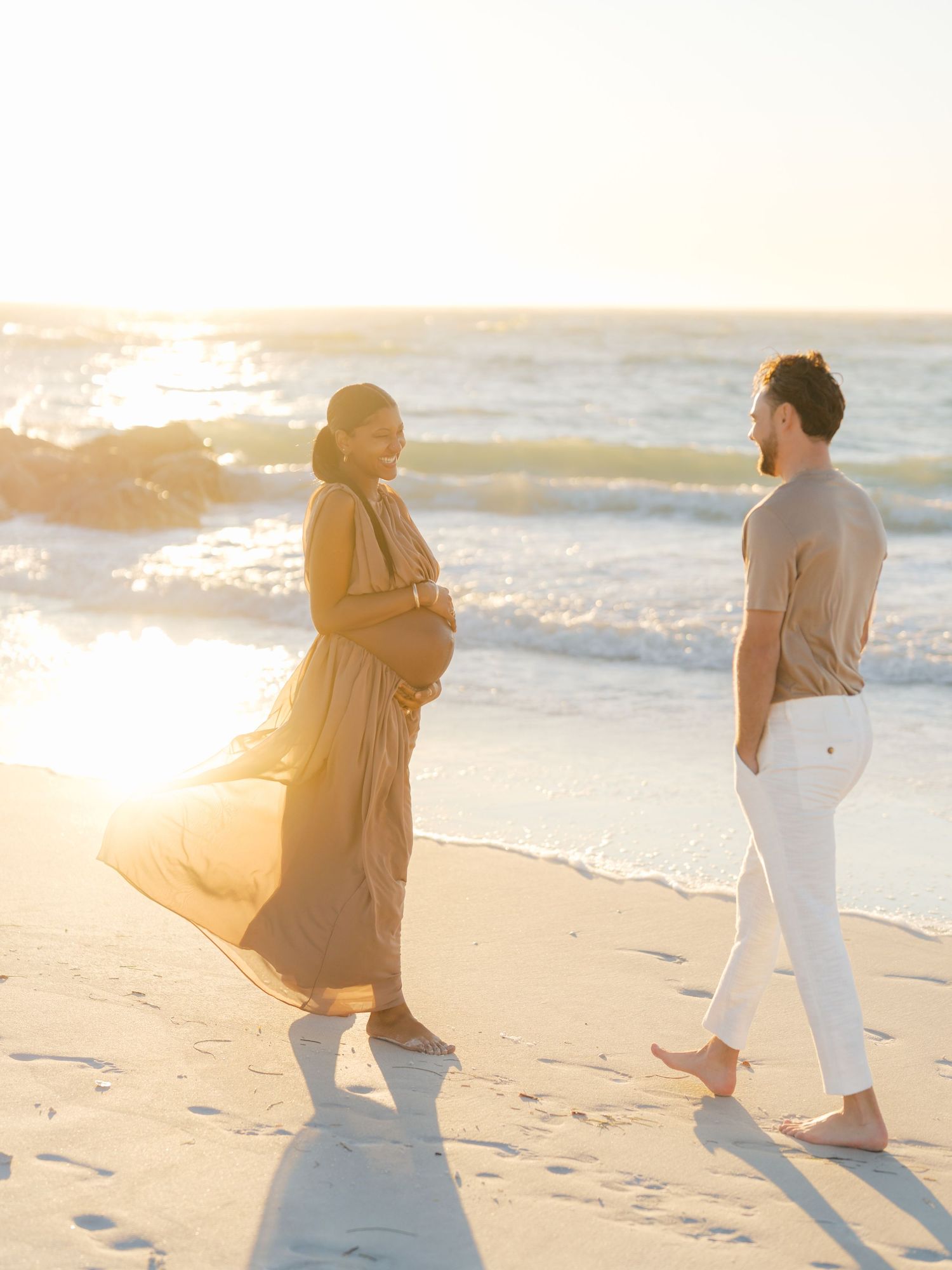 Pregnant couple shares a tender moment on a sunlit beach at golden hour, waves crashing behind them.
