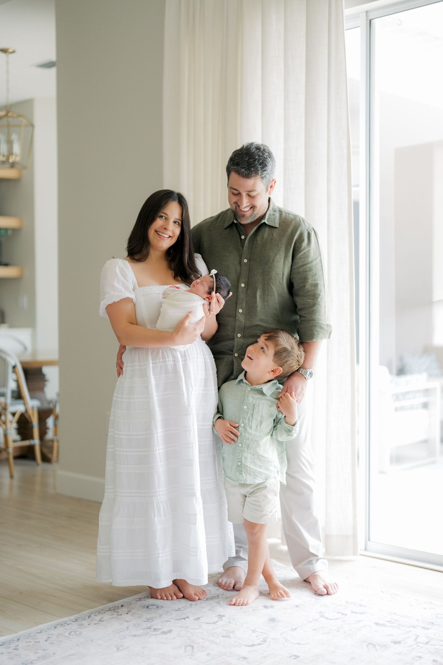 A happy family of four poses indoors, mother in white dress holding newborn, father and toddler son smiling together.
