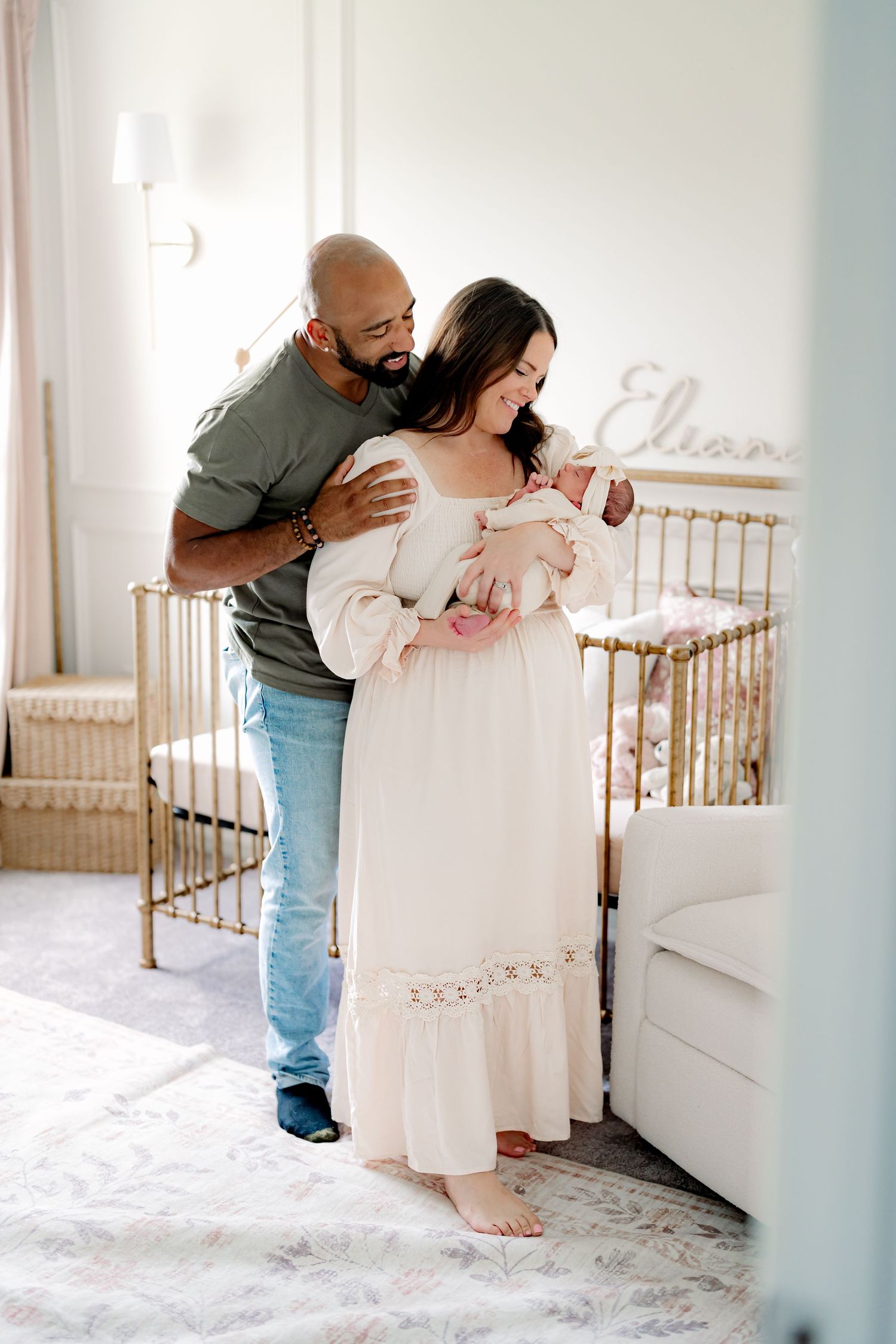 Parents admiring newborn baby in nursery with gold crib in background.