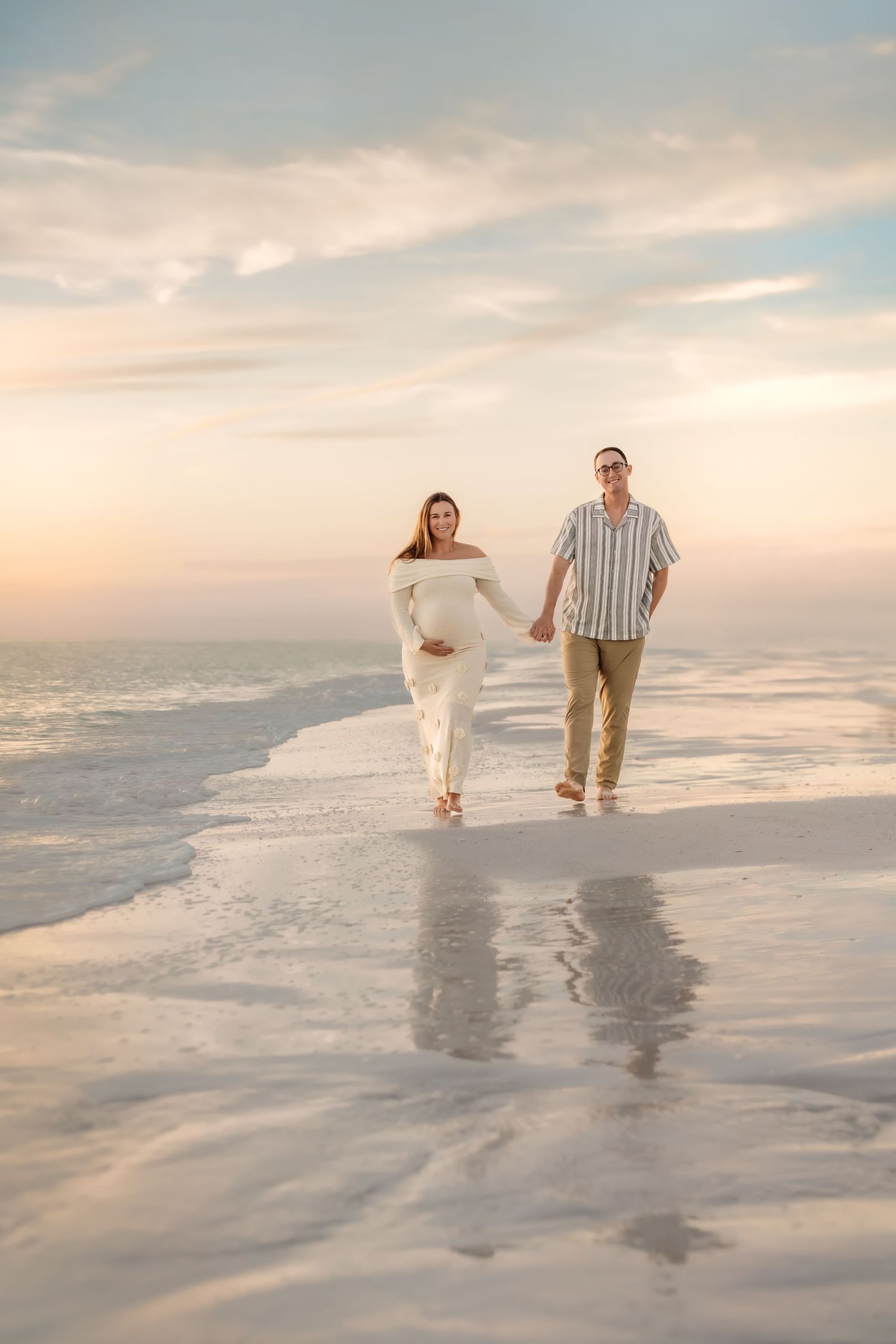 A couple walks along a misty beach at sunset, leaving footprints in the wet sand as waves roll in.