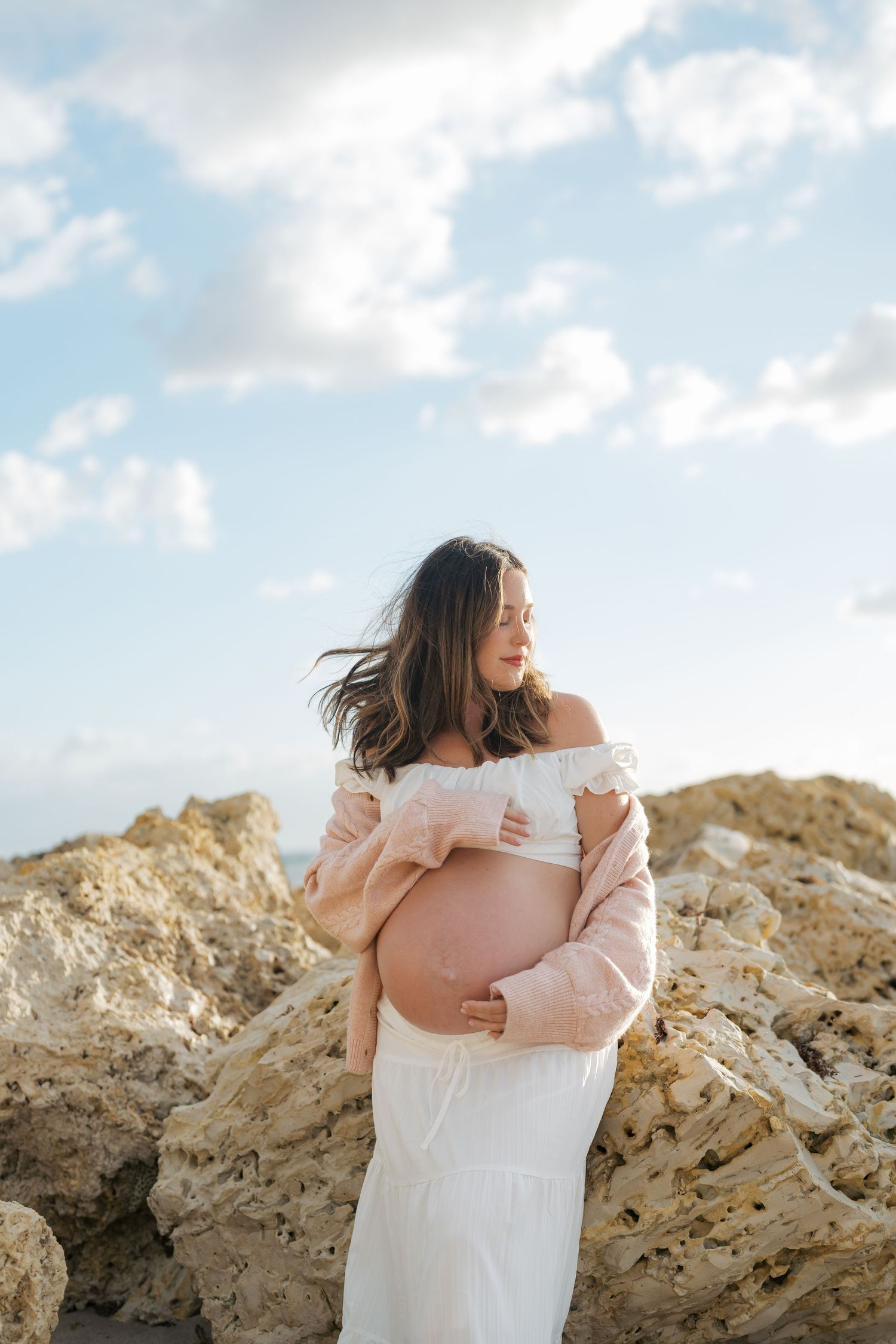 Pregnant woman in white dress and pink jacket cradling baby bump on rocky beach with cloudy sky.