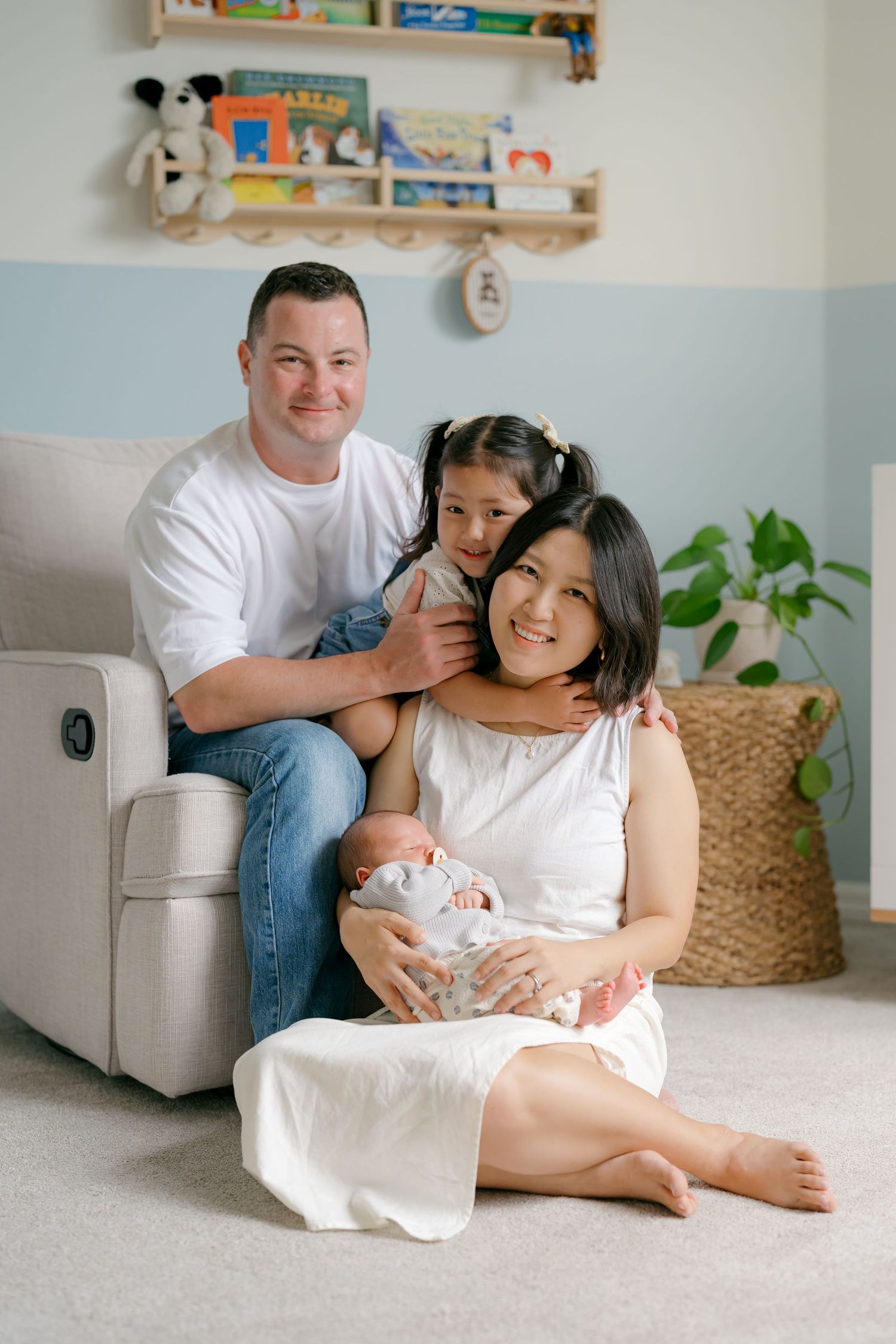 Happy family of four posing in a cozy nursery, parents smiling with toddler and newborn baby in a light blue room.