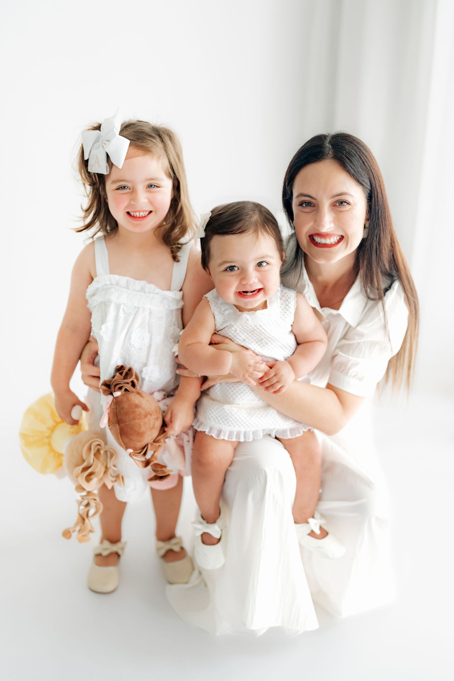 Three joyful children in white dresses sit together and laugh while playing in a bright studio setting.