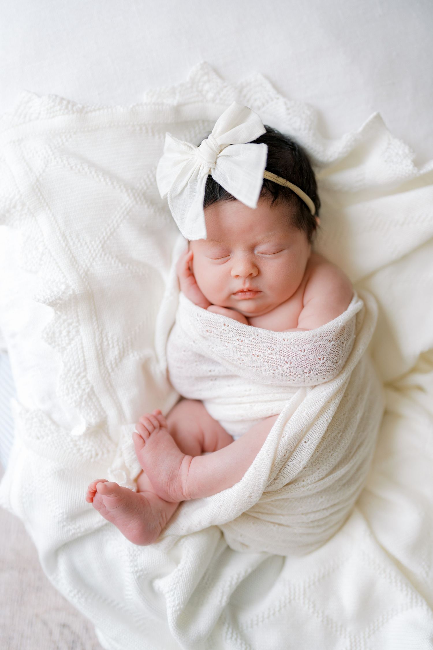 Sleeping newborn baby girl swaddled in white wrap with bow headband on white blanket.