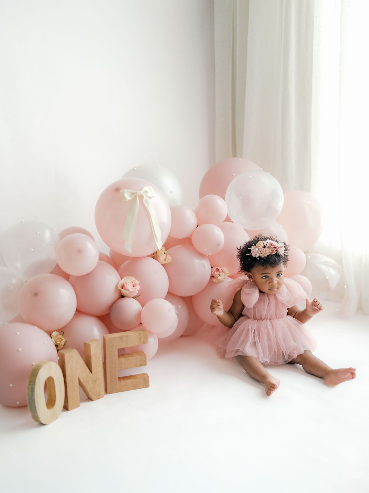 A milestone photo collage featuring a baby in pink dress with gold 'ONE' letters and blush balloons against white backdrop.