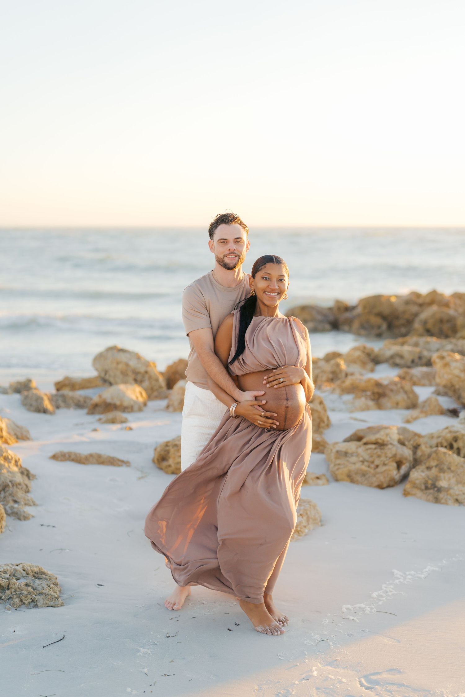 Couple embracing on beach at sunset, man holding pregnant womans belly in flowing tan dress.