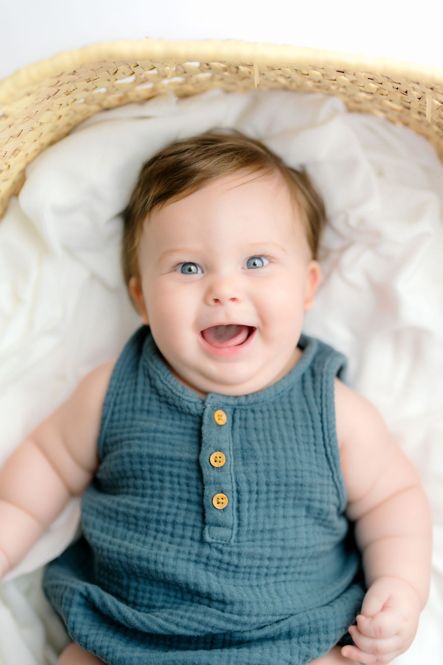 A happy baby wearing a blue sleeveless romper with wooden buttons lies on white bedding in a woven basket.