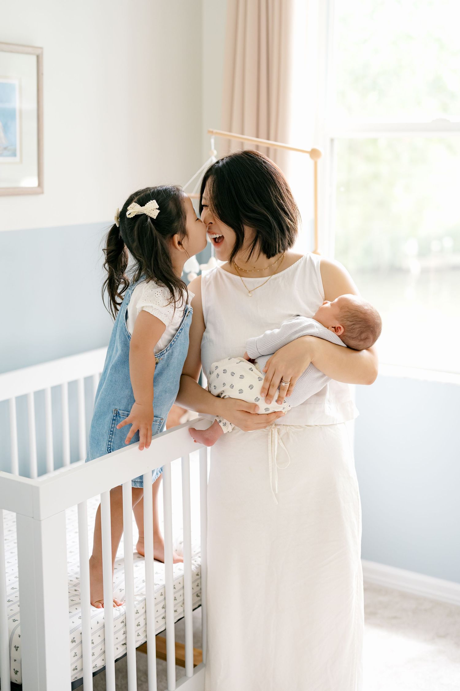 A mother in white holds her newborn while her toddler daughter stands in a crib, sharing a joyful nose-to-nose moment.