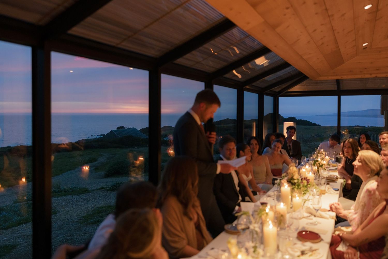 Elegant seaside dinner party at dusk with guests seated at a long candlelit table in a glass-walled dining room.