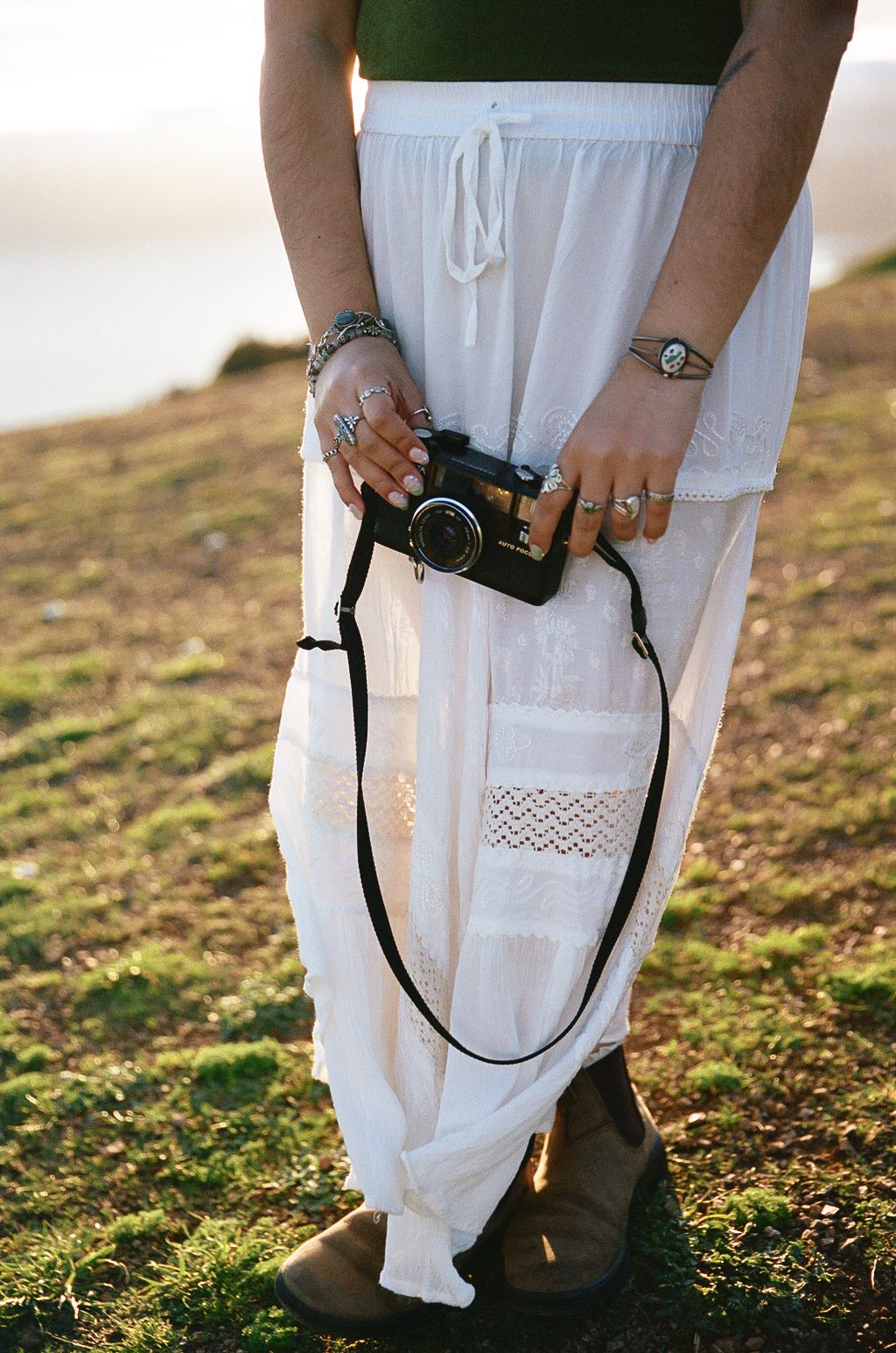 Someone wearing white pants holds a vintage camera with rings and bracelets visible against a sunset background.