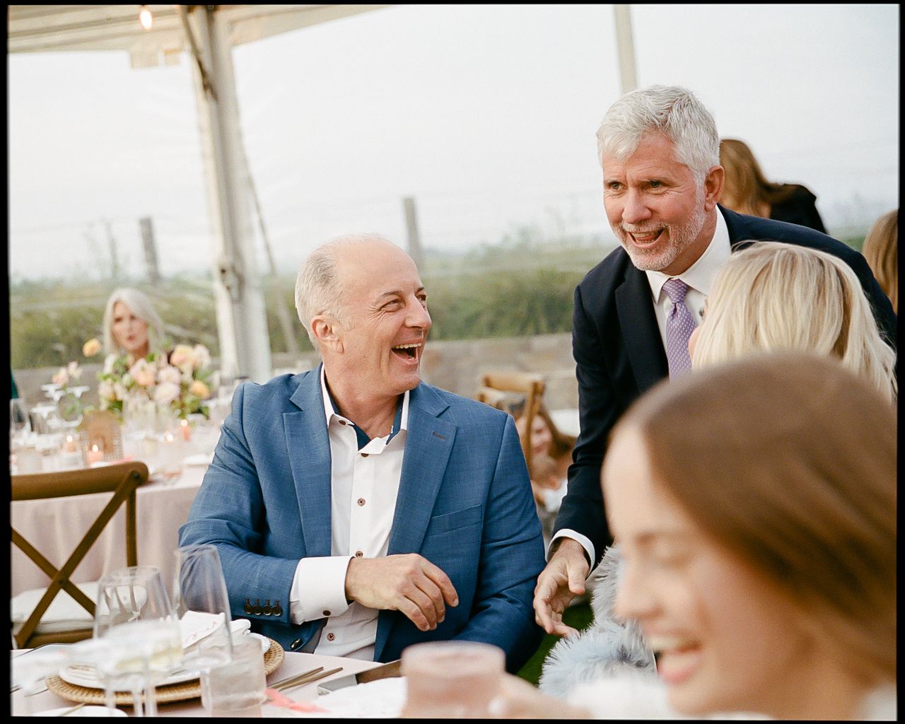 Men in suits laughing and socializing at an elegant outdoor wedding reception dinner under a tent.