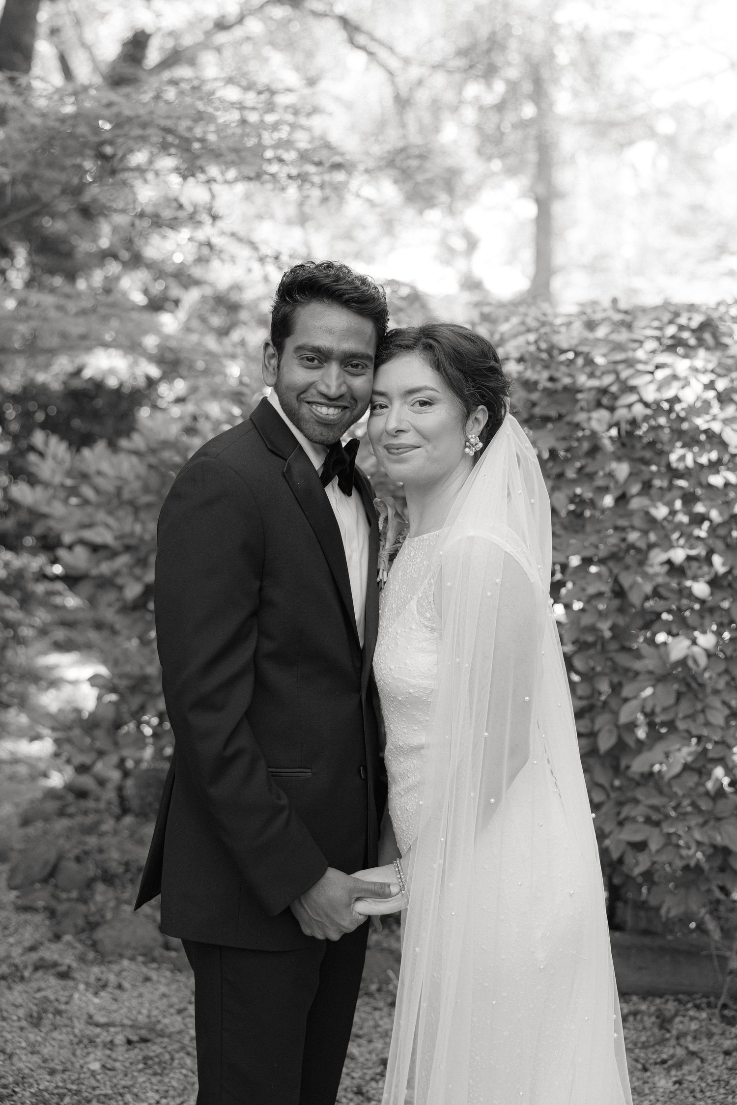 Black and white wedding portrait of bride in veil and groom in tuxedo posing together outdoors.