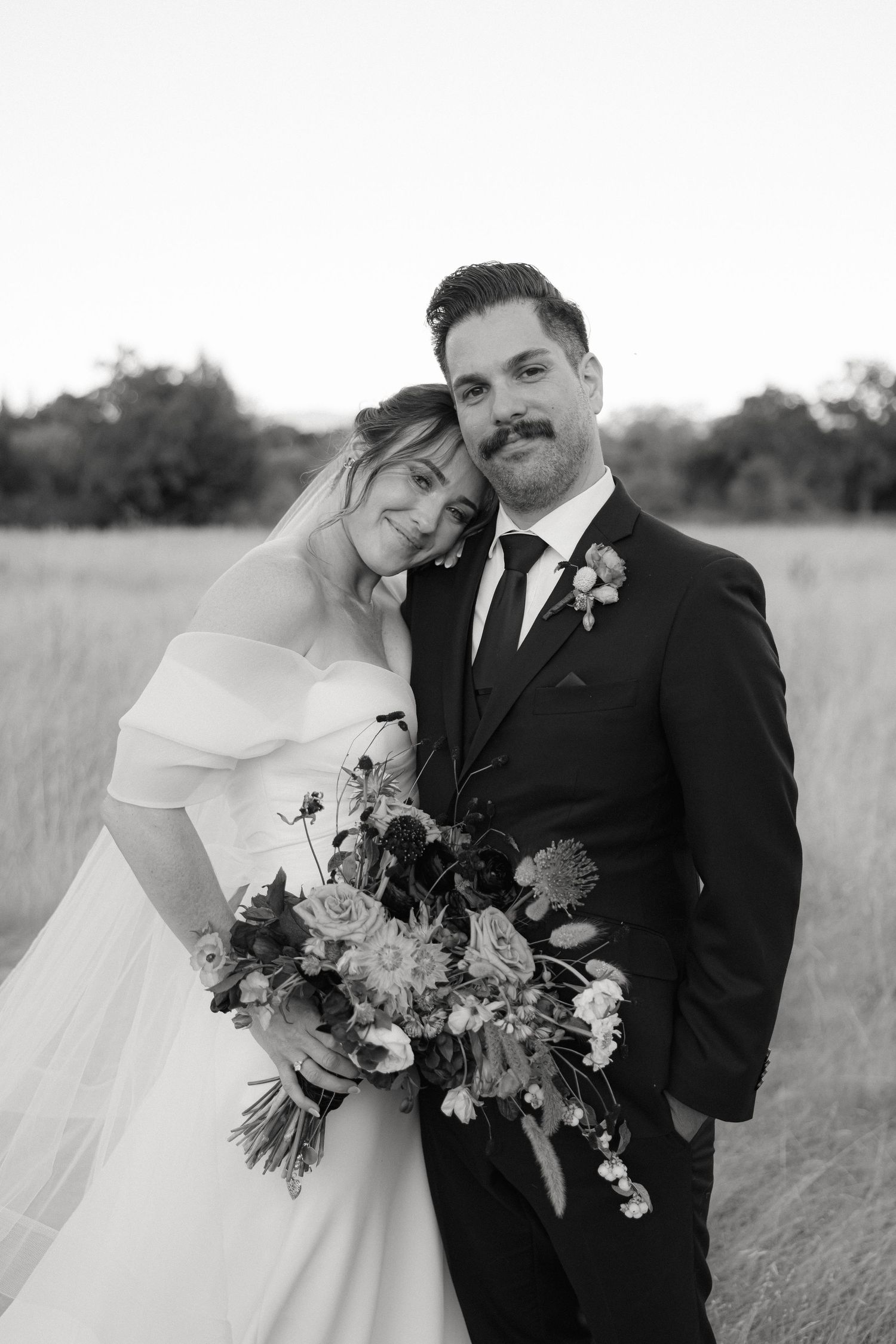 A romantic black and white portrait of newlyweds with a floral bouquet in a field.