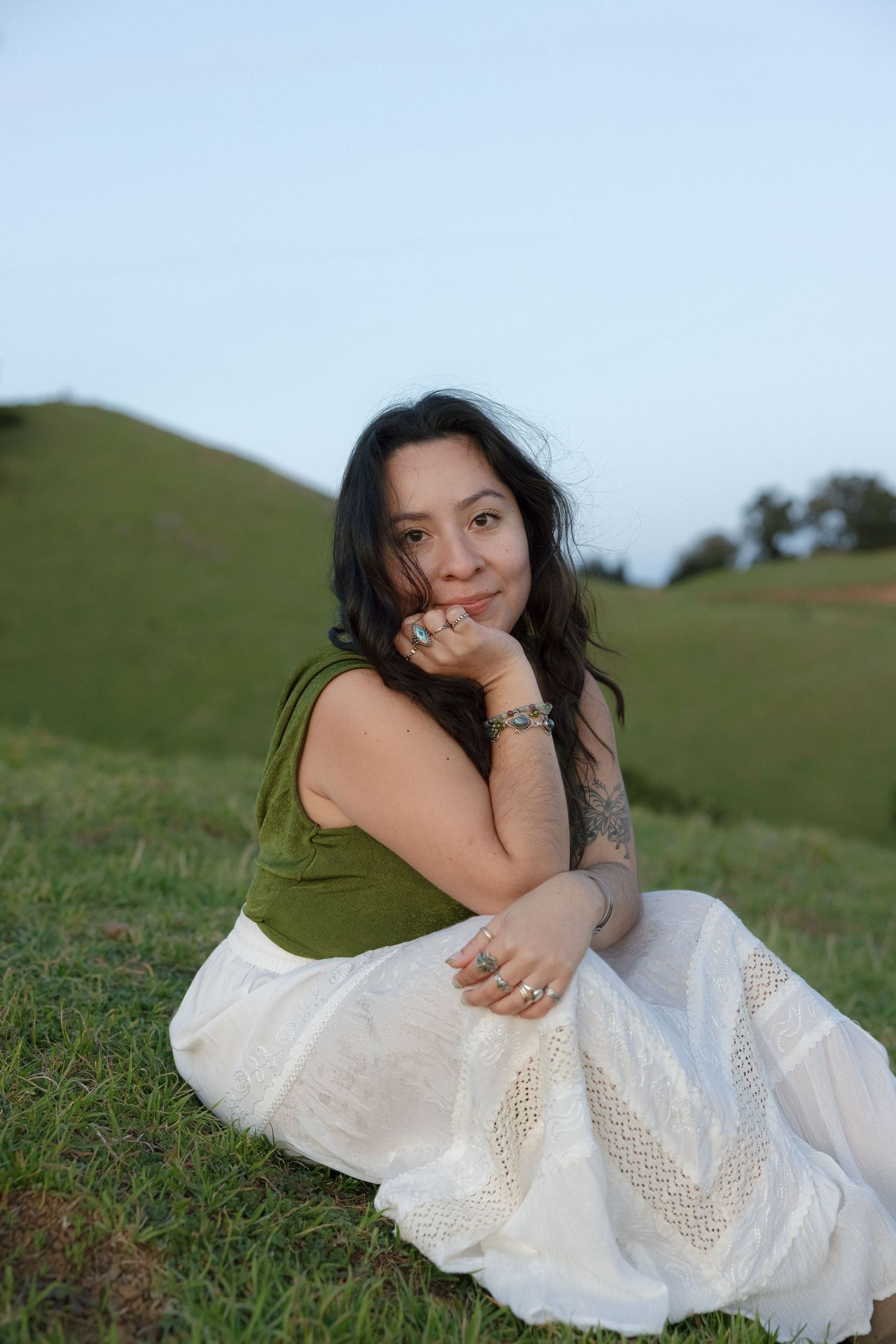 A person in a white lace dress and olive top poses gracefully on grassy hillside during golden hour.