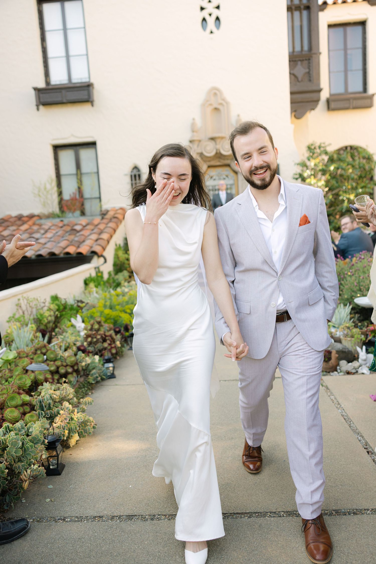 A couple in white attire share a joyful moment on their wedding day in a garden setting.