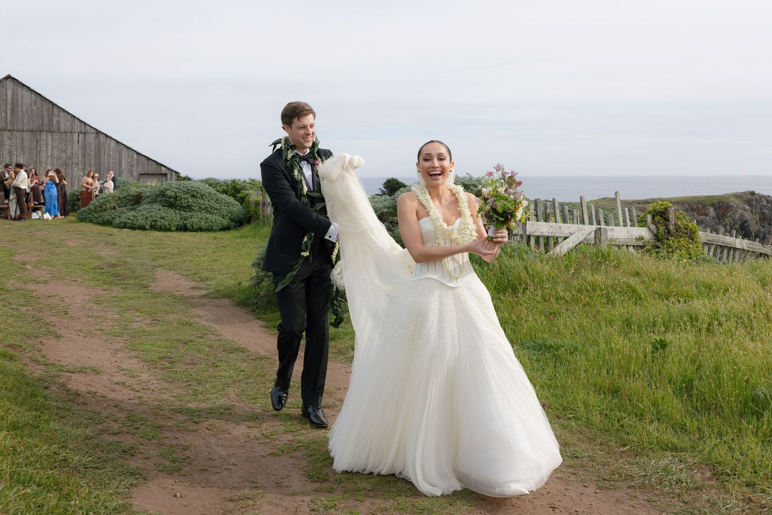 Bride and groom walking outdoors on a coastal path, smiling, bride holding bouquet and dress, ocean view background.