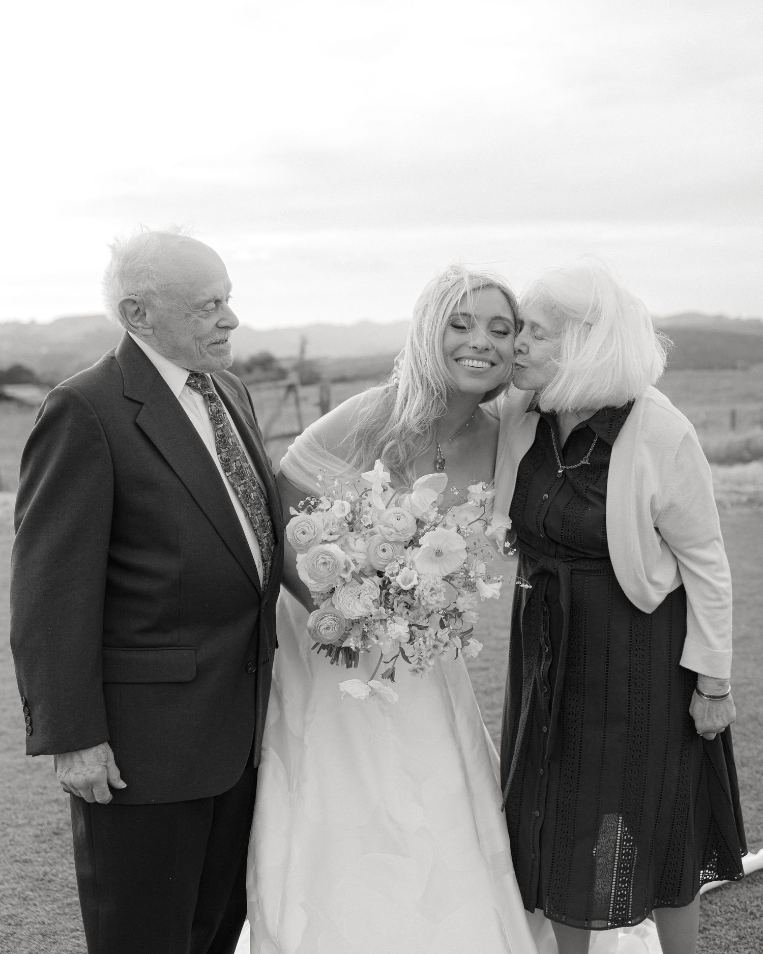 Black and white photo of a bride holding bouquet receiving a kiss on cheek from elderly woman beside suited older man.