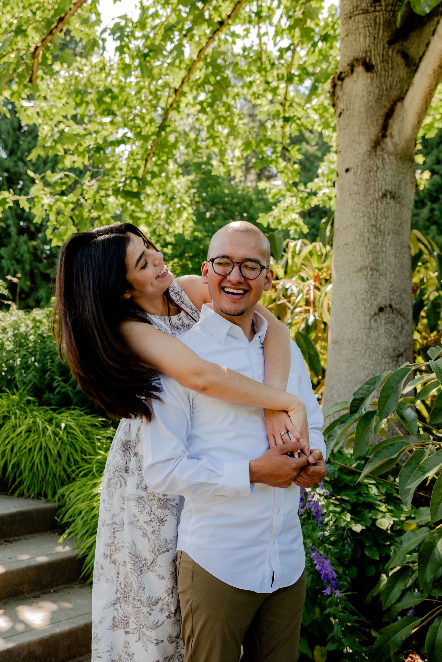 Happy couple embracing and laughing together in a lush green garden surrounded by trees and flowers.