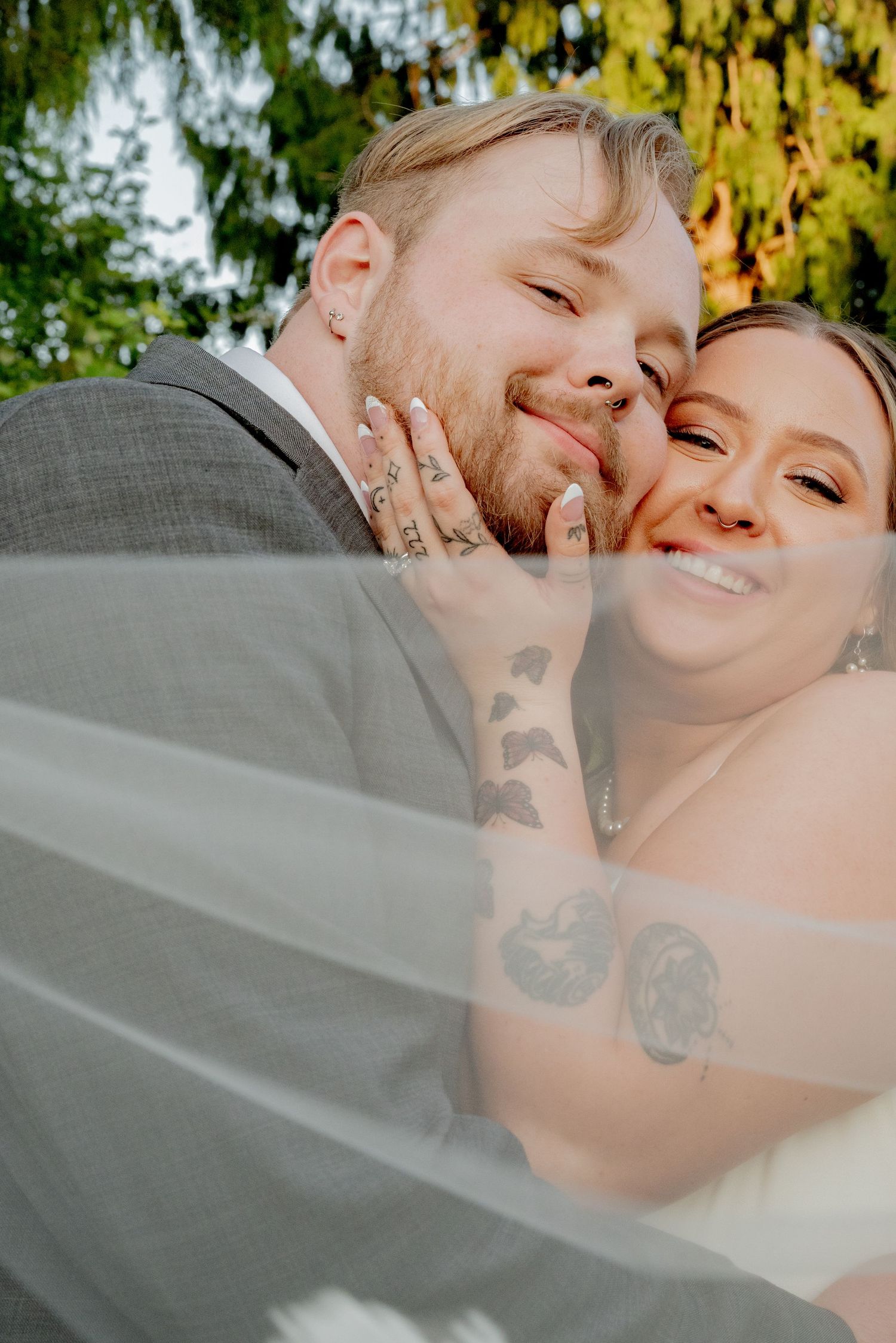 Arlignton, Washington, A joyful wedding couple embraces under a veil while showing off tattoos and rings in an outdoor setting.