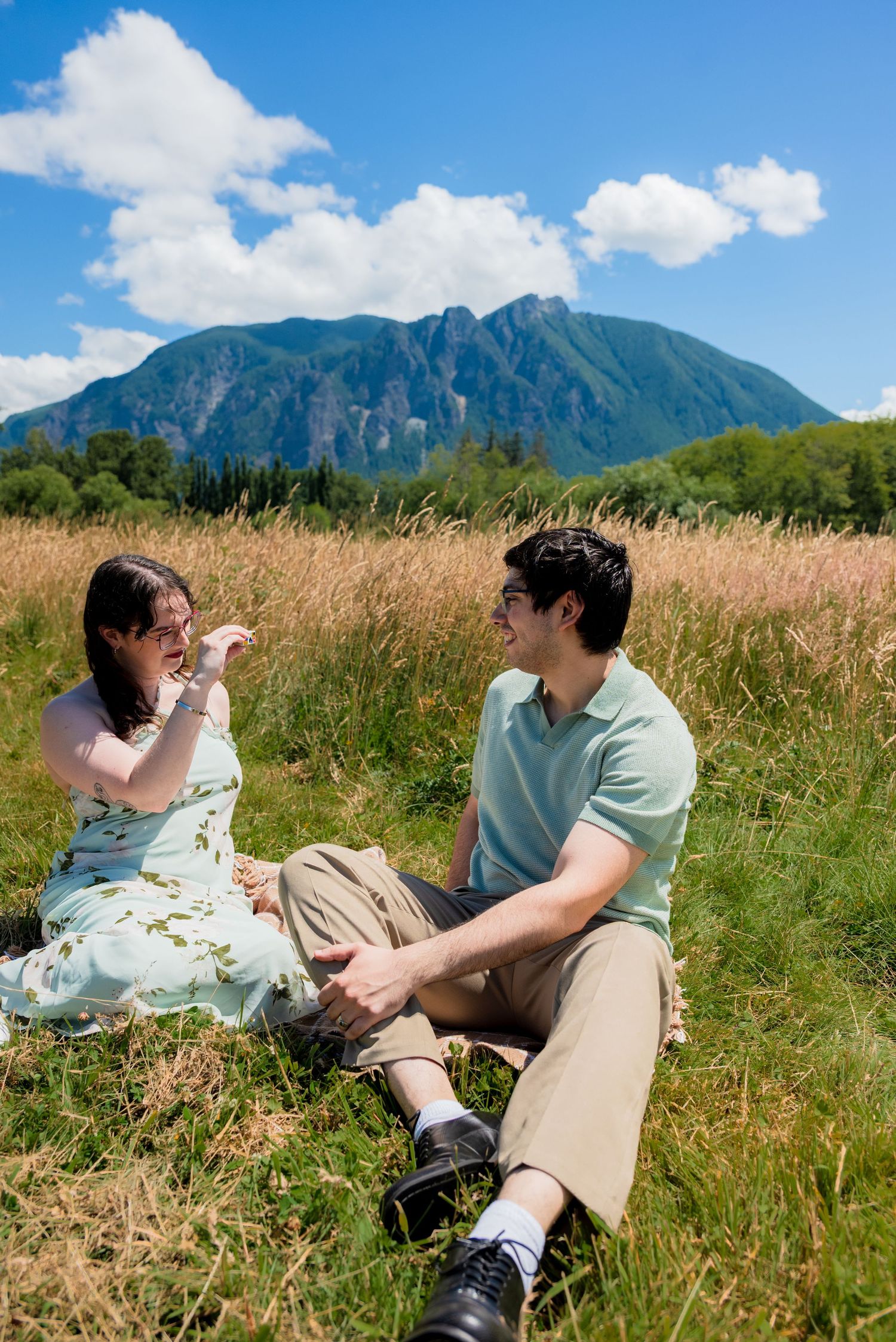 Couple sitting together in a grassy field laughing, with a dramatic mountain view behind them.