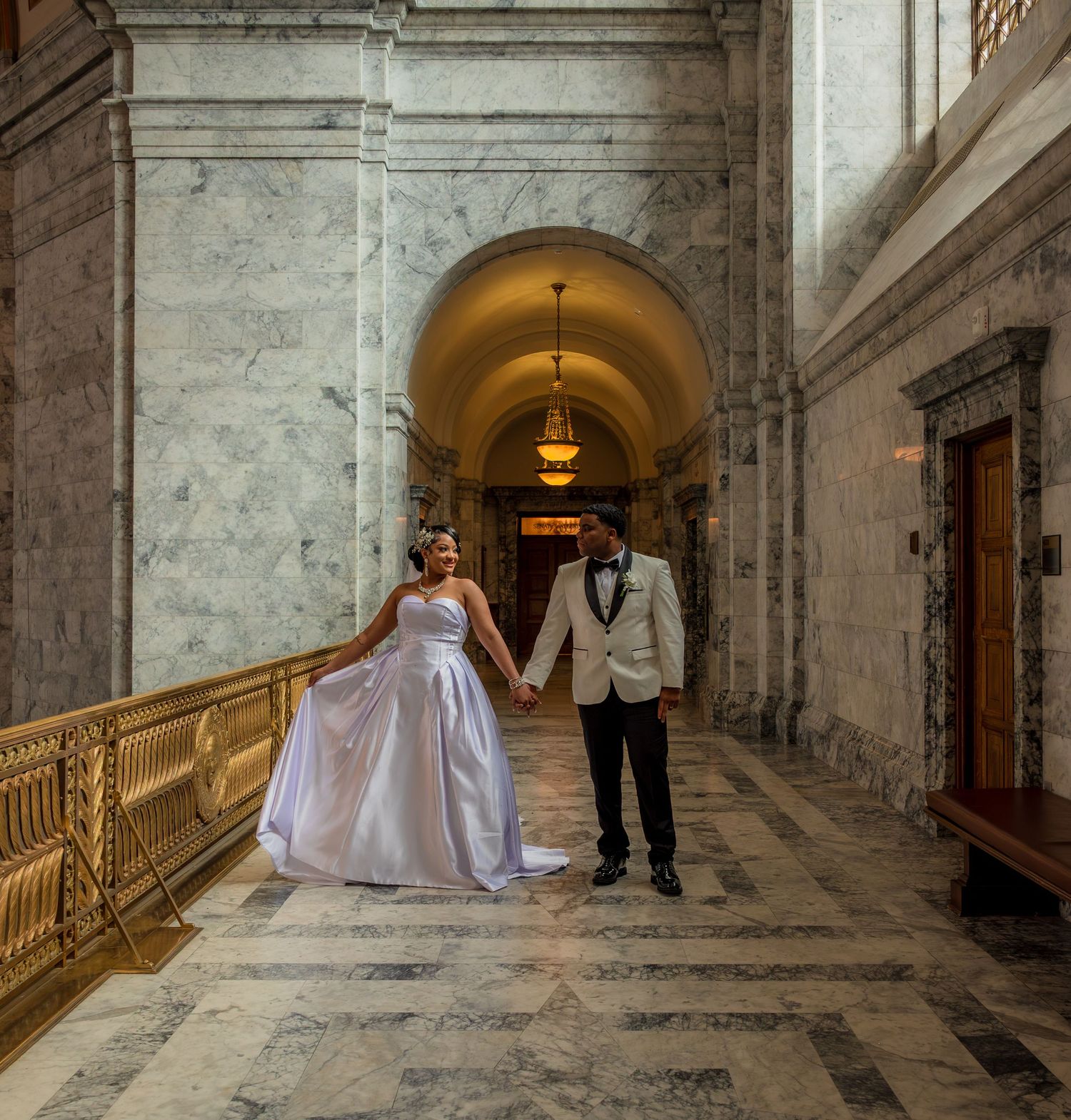 Bride and groom hold hands in an elegant marble hallway with arched ceilings and ornate gold railings.