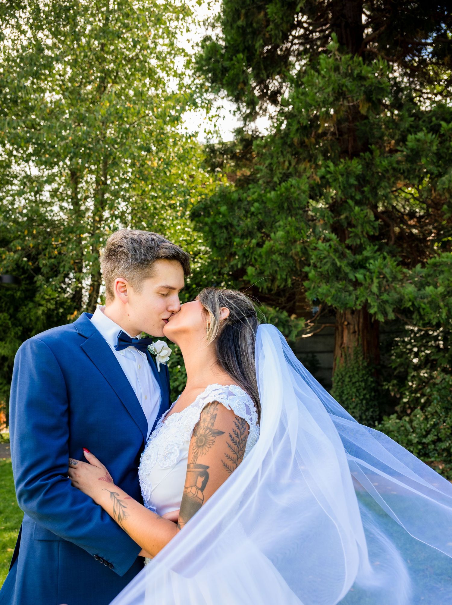 Newlyweds kissing in a garden, groom in blue suit, bride in lace dress with flowing veil billowing.