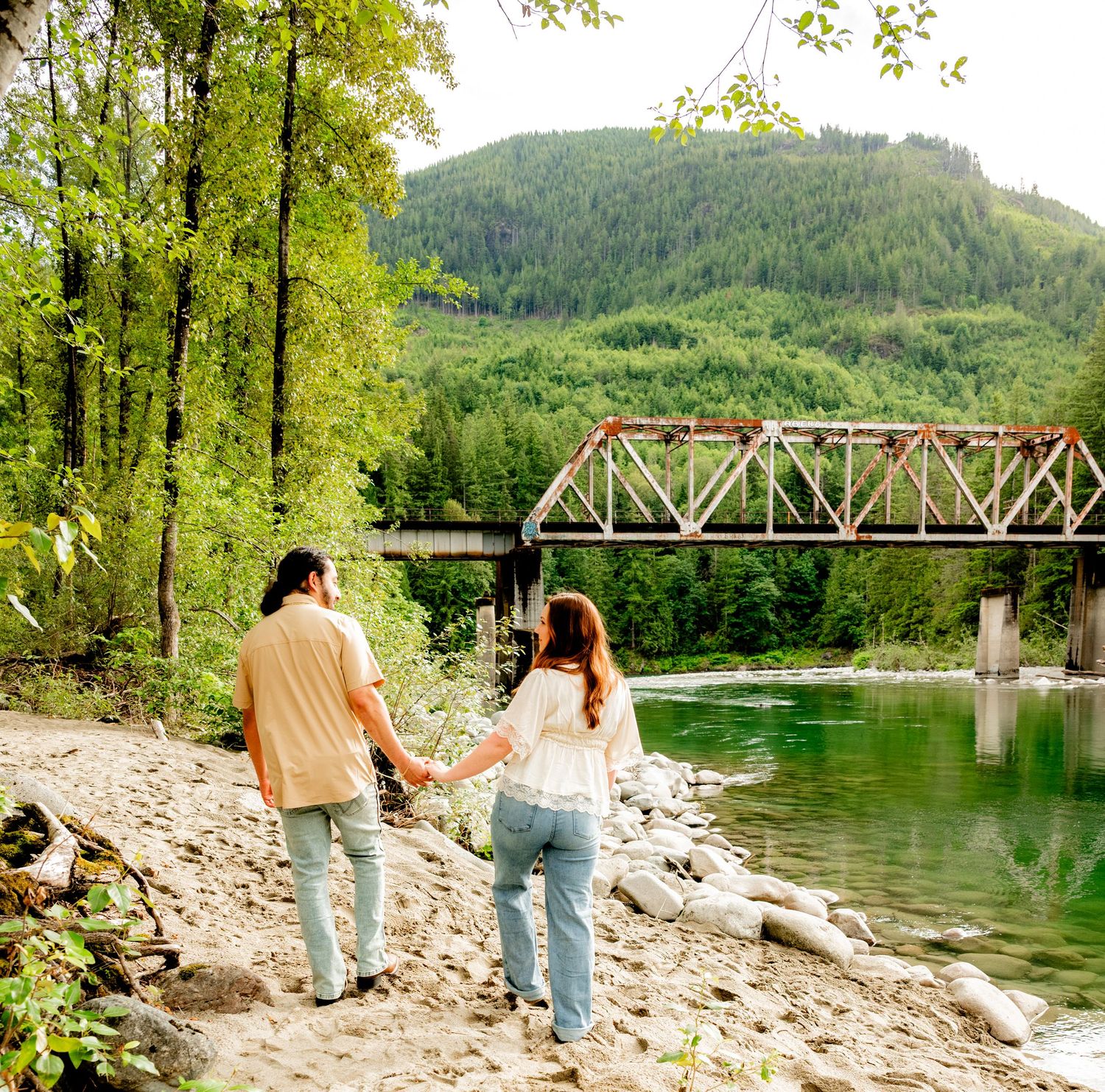 Couple walks hand in hand along a rocky riverbank with a historic iron bridge and green mountains.