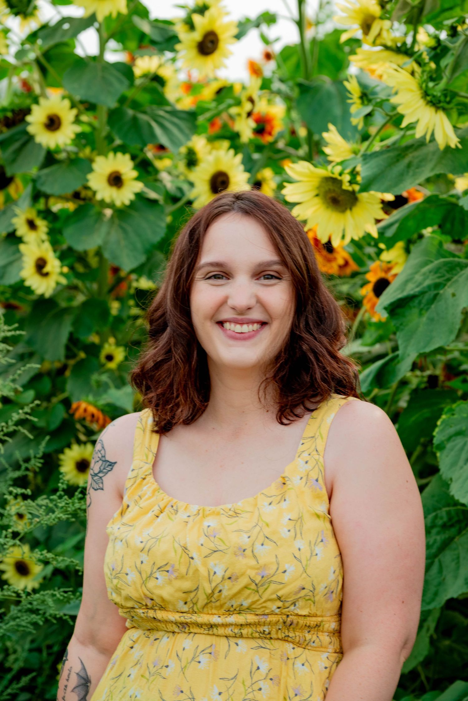 A cheerful portrait in a yellow floral dress against vibrant blooming sunflowers and green foliage in a garden setting.