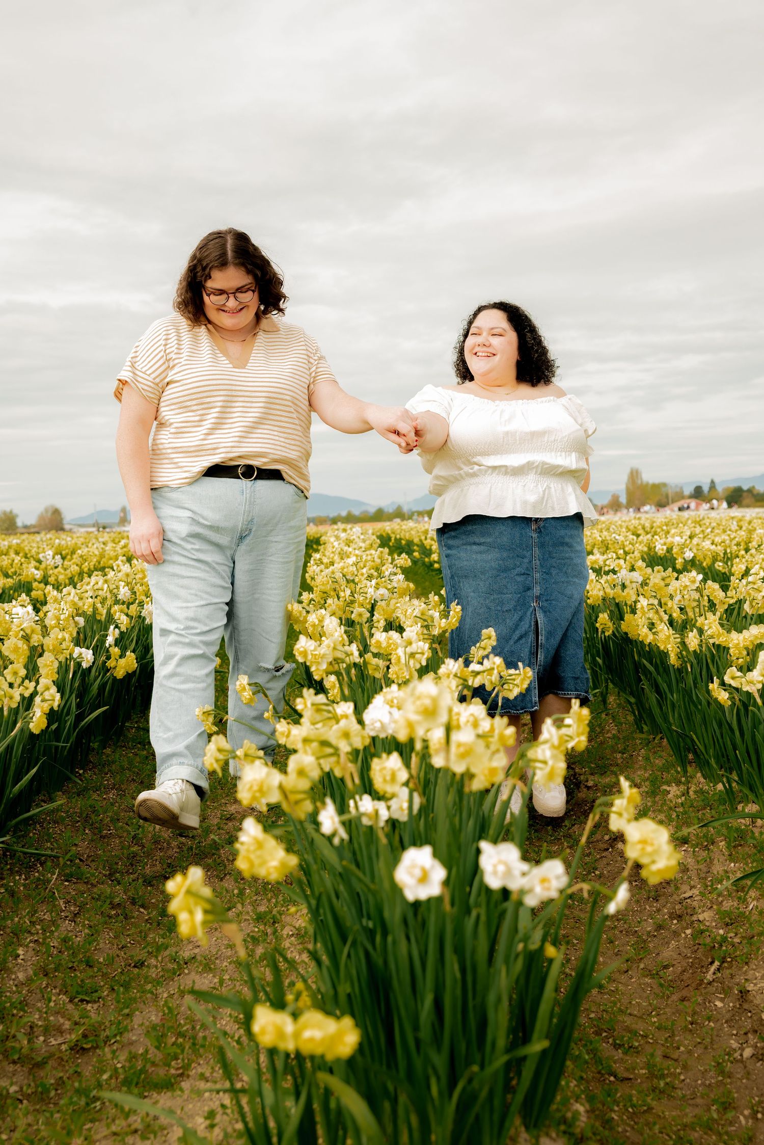 Two women holding hands and walking through a vibrant yellow daffodil field under a cloudy sky.