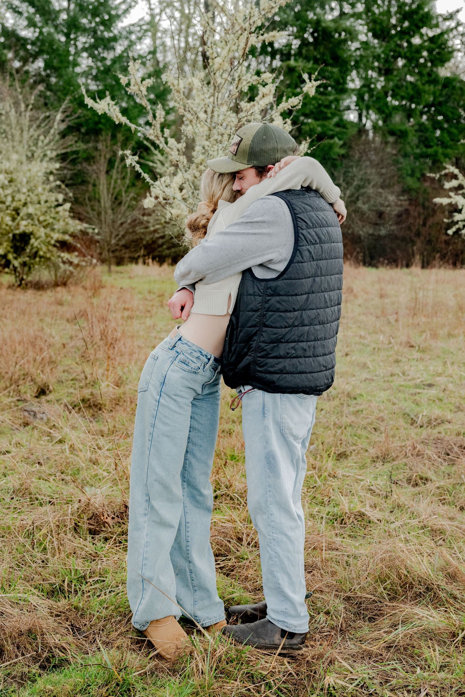 Olympia, Washington, Two people embrace in a tender moment while standing in a grassy field with evergreen trees in the background.