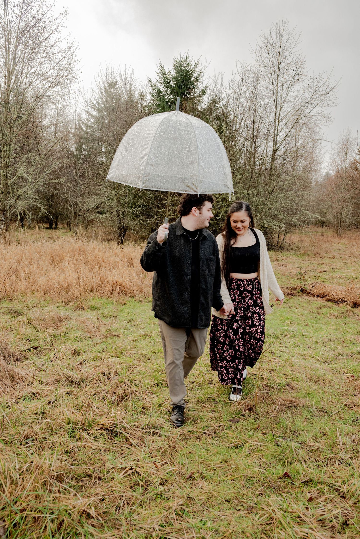 Olympia, Washington, A couple walks together under a clear umbrella on a misty day in a grassy field surrounded by bare trees.