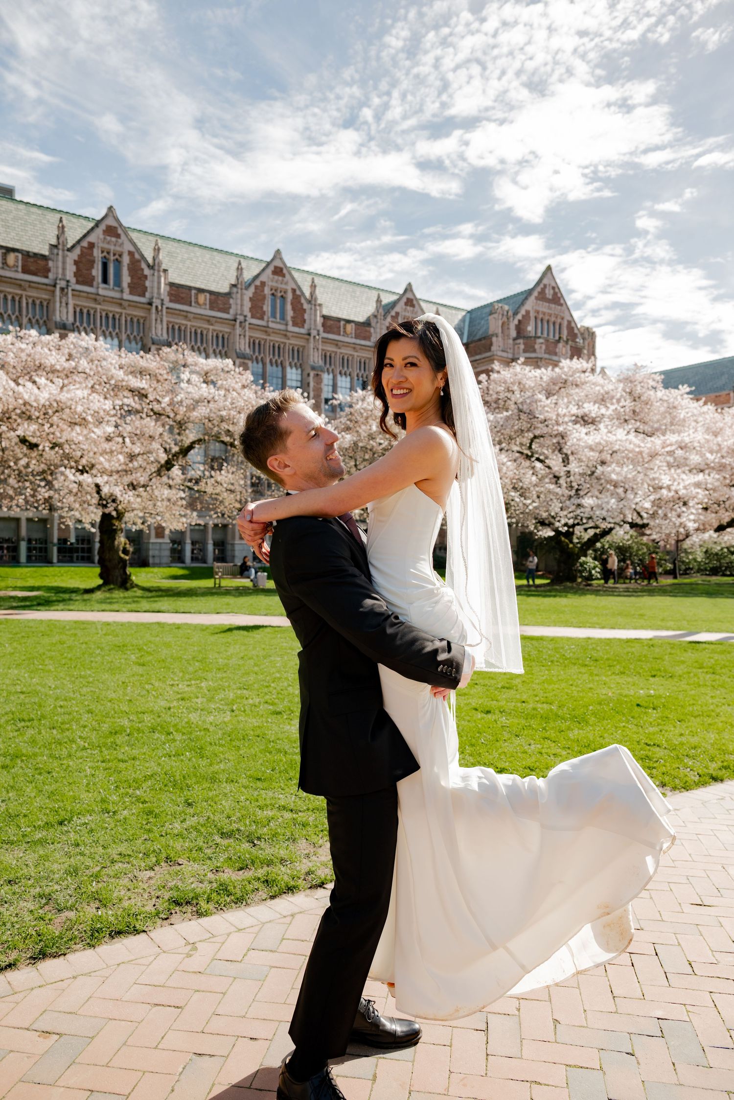 Groom lifting bride in white gown among cherry blossoms at University of Washington on their wedding day.