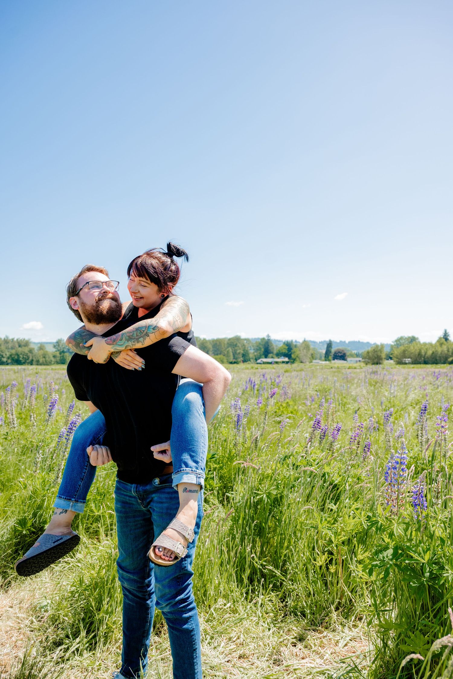 Bearded man giving his partner a piggyback ride in a blooming purple lupine field under blue sky.