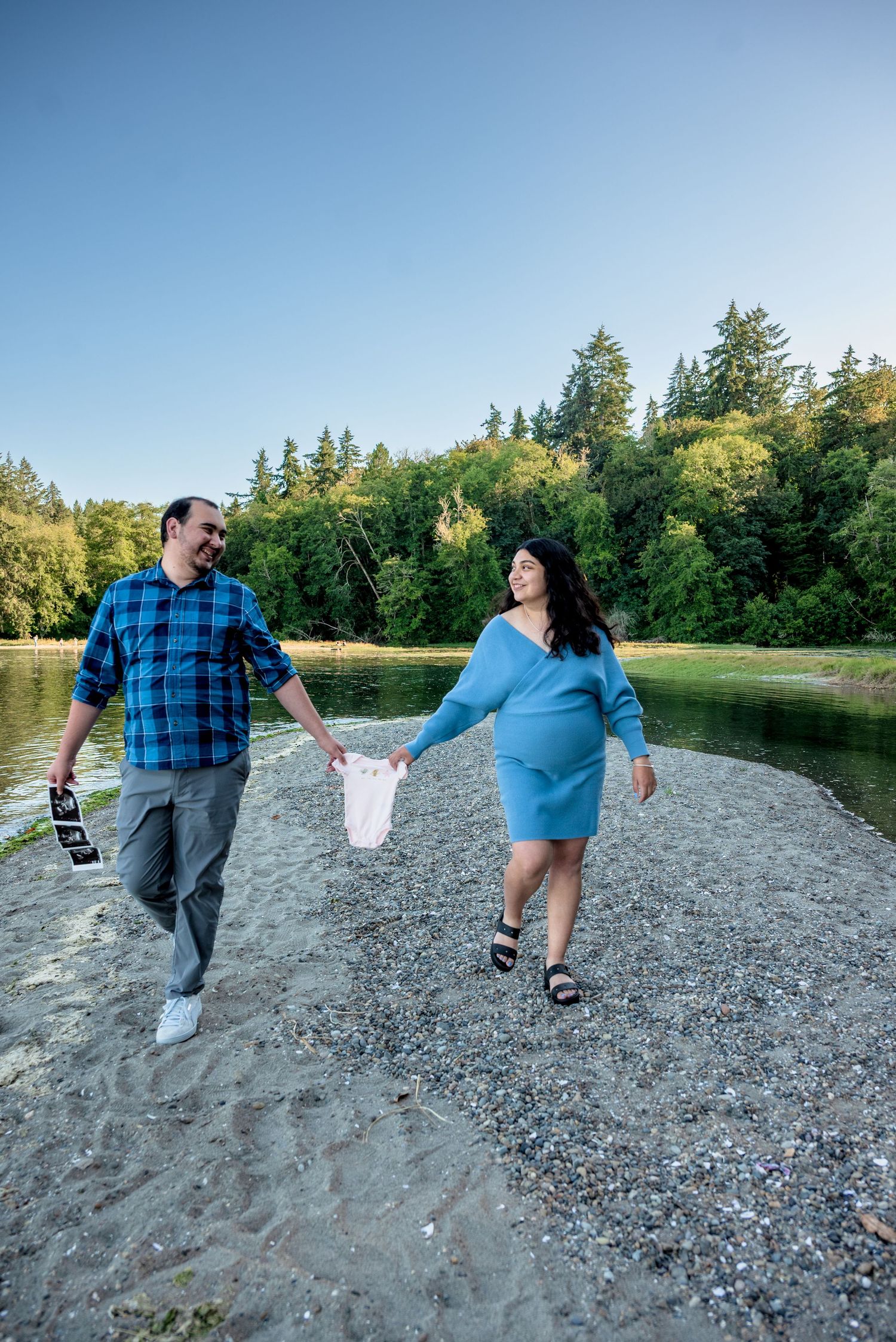 Pregnant couple holding hands and walking along a sandy riverbank with lush green trees in the background.