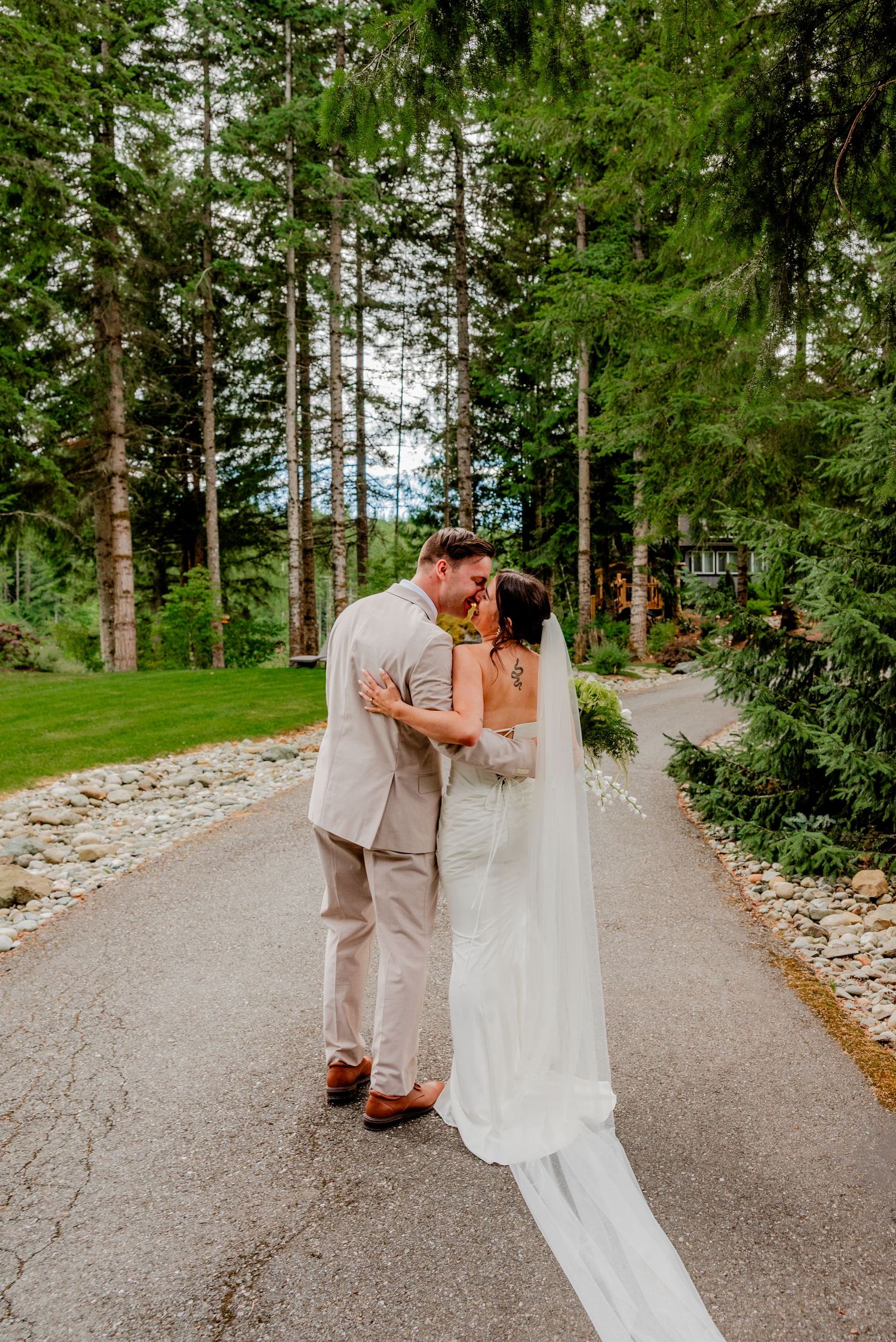 Newlyweds share a kiss on a tree-lined path, bride wearing a long veil in a serene forest setting.