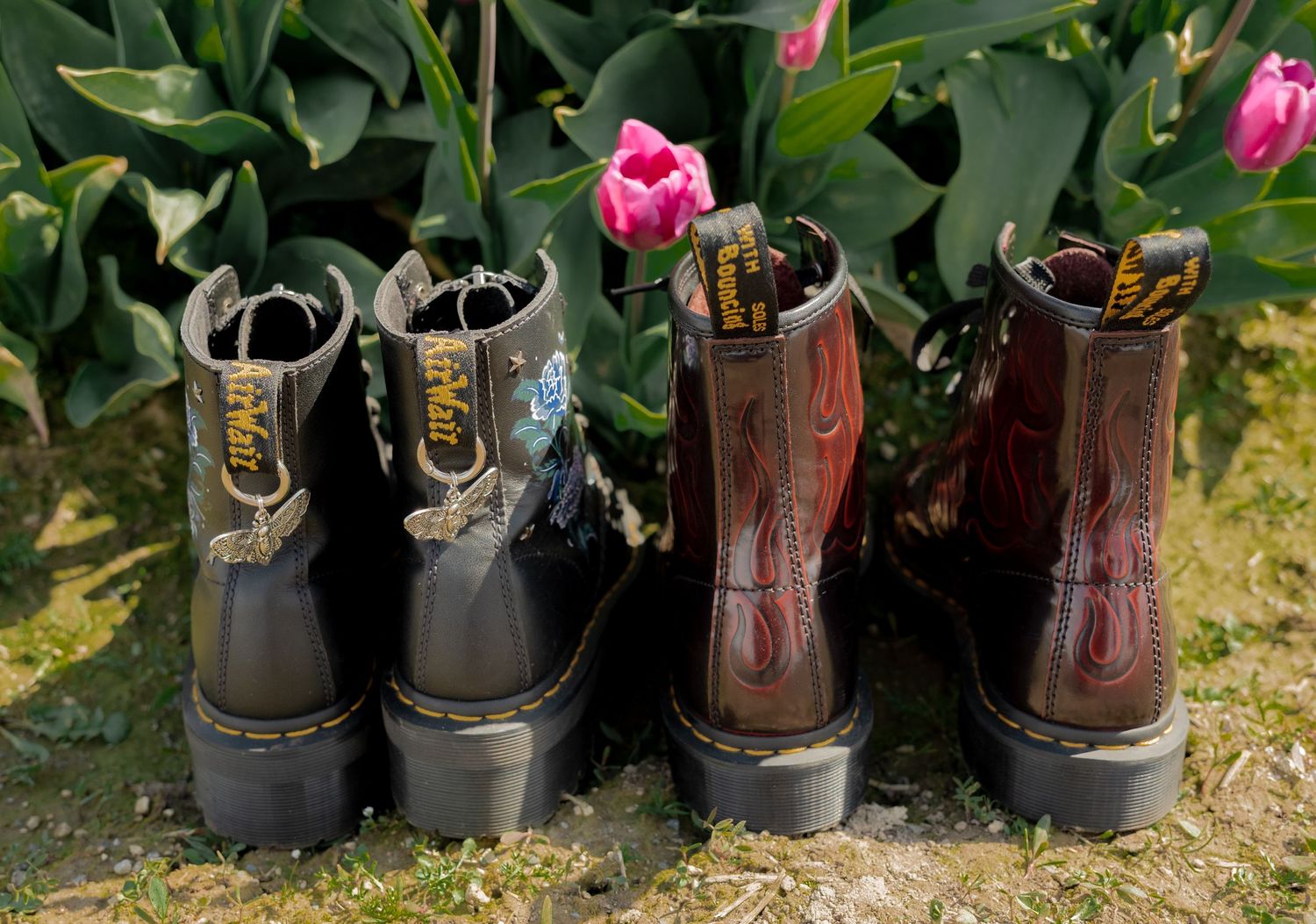 Two pairs of combat boots, one black and one brown, displayed upright among green tulip stems with pink blooms.